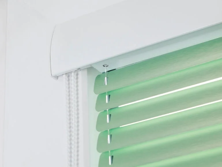 Nahaufnahme eines weißen Rollos mit grünen Lamellen an einem Fenster, das teilweise geschlossen ist.