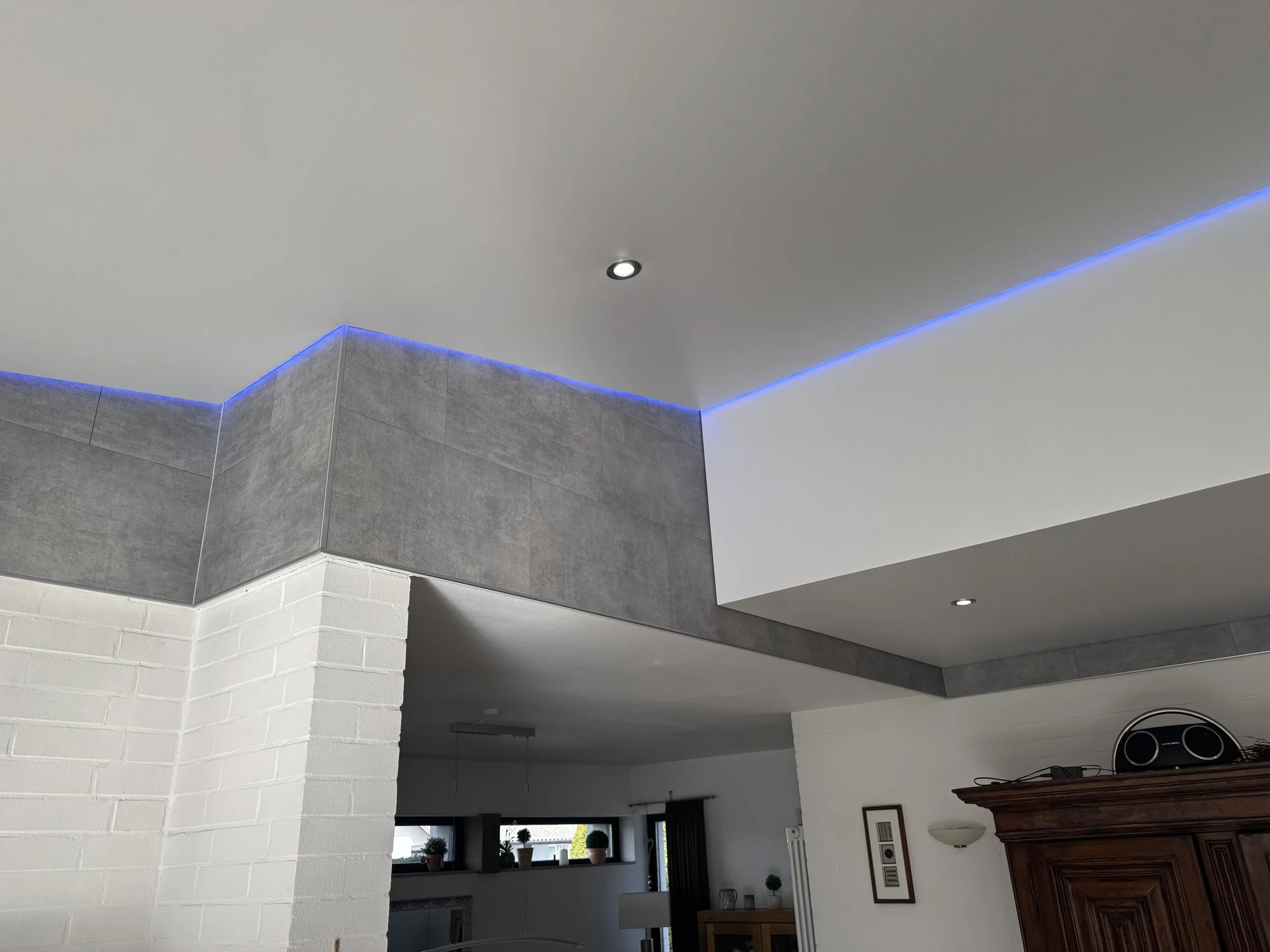 Innenaufnahme eines Raums mit weißem Decke, grauen Fliesen an den Wänden, blauer LED-Beleuchtung an der Decke und einem Fenster mit Pflanzen davor. Im unteren rechten Bereich ist ein alterlege Möbelstück zu sehen.