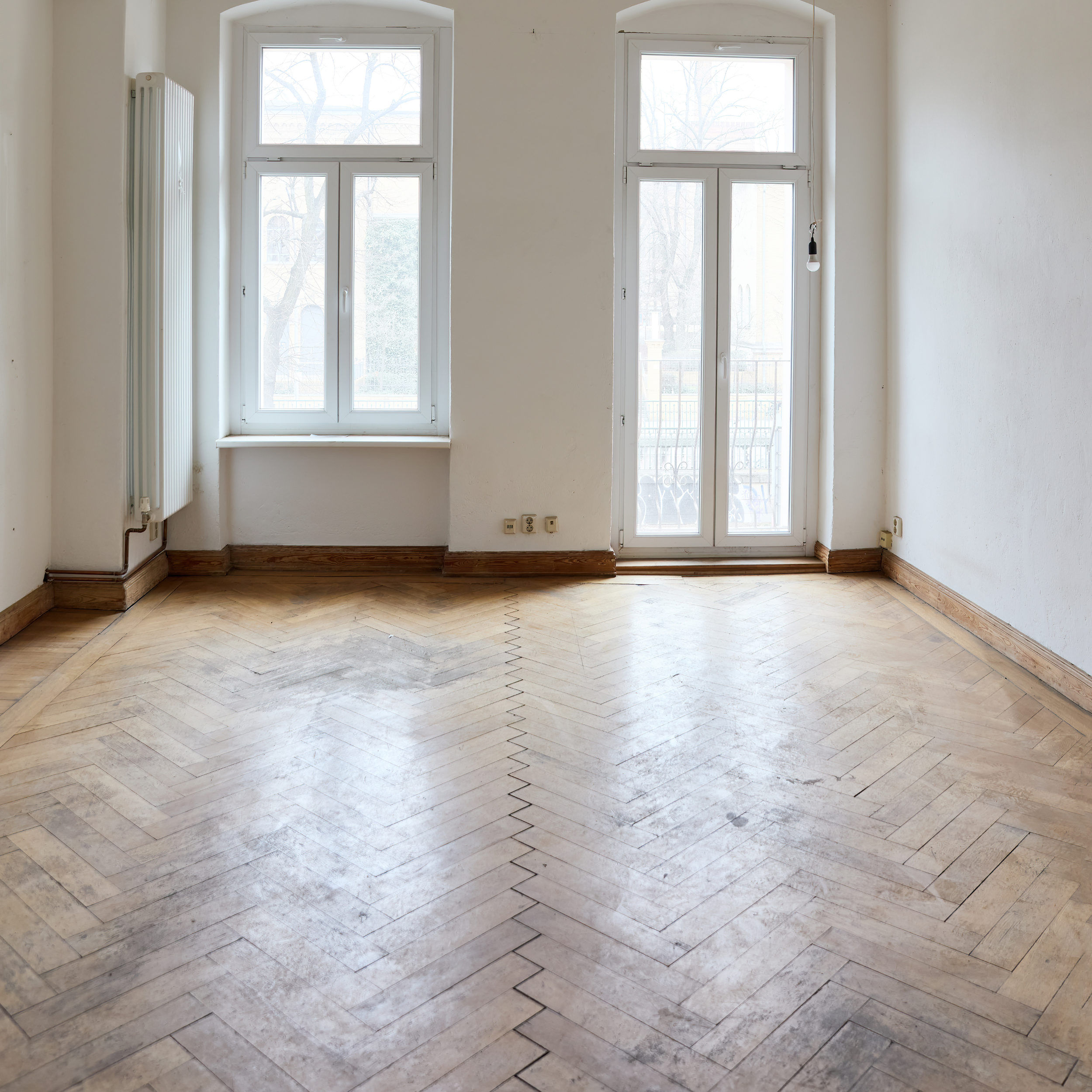 Ein heller Raum mit großen Fenstern, die einen Blick auf Bäume im Winter freigeben. Der Boden ist aus Holz im Fischgrätmuster, die Wände sind weiß, und es gibt eine Heizkörper an der linken Wand, sowie Stromanschlüsse nahe dem Boden.