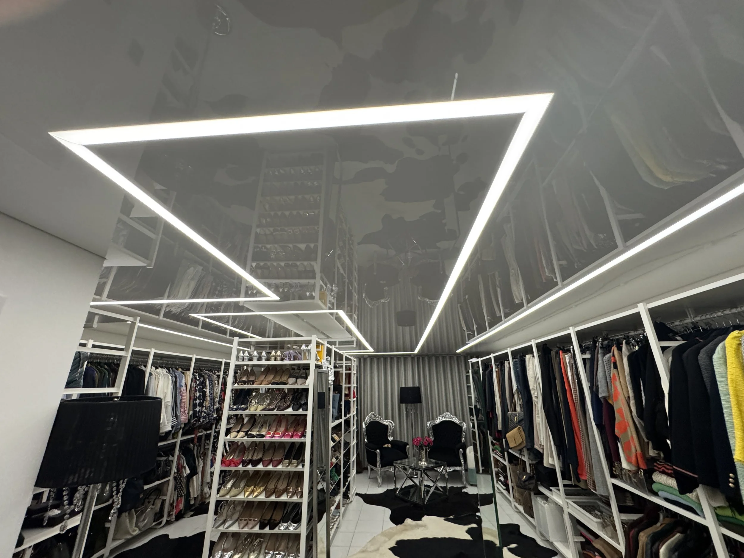 Innenseite eines Modegeschäfts mit mehreren Regalen voller Schuhe und Kleidung, moderne LED-Beleuchtung an der Decke, einige Stühle und ein kleines Tisch in der Mitte, dekorative Spiegelstühle in der Ecke.