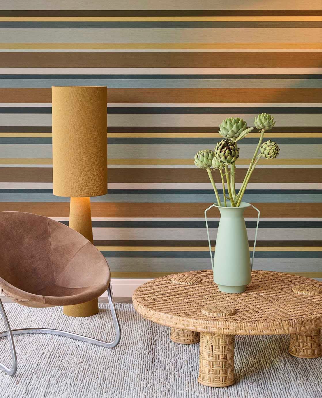 Ein Wohnzimmer mit gestrichener Wand mit horizontalen Streifen in Braun, Grau, Taupe, Hellblau und Creme. Es gibt einen runden Tisch aus geflochtenem Holz mit einem grünen Vasen mit Kunstpflanzen, neben einem braunen Sessel und einer Stehlampe in Gra