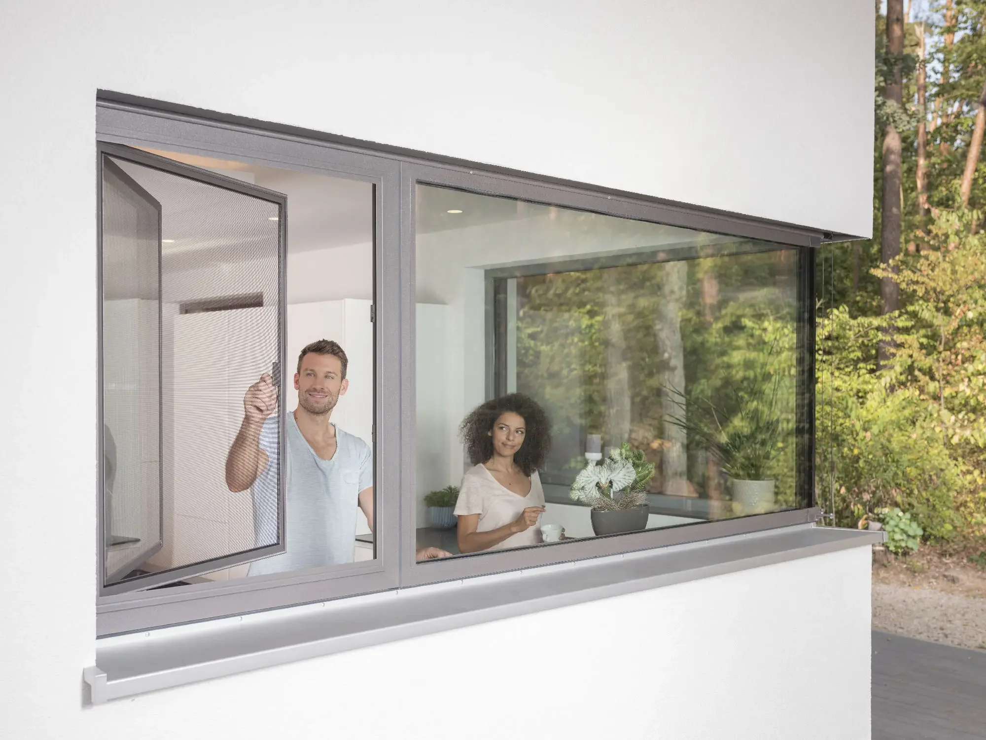 Drei Menschen sehen durch ein großes Küchenfenster, eines trinkt Kaffee, das andere hält eine Pflanze, draußen sind Bäume sichtbar.