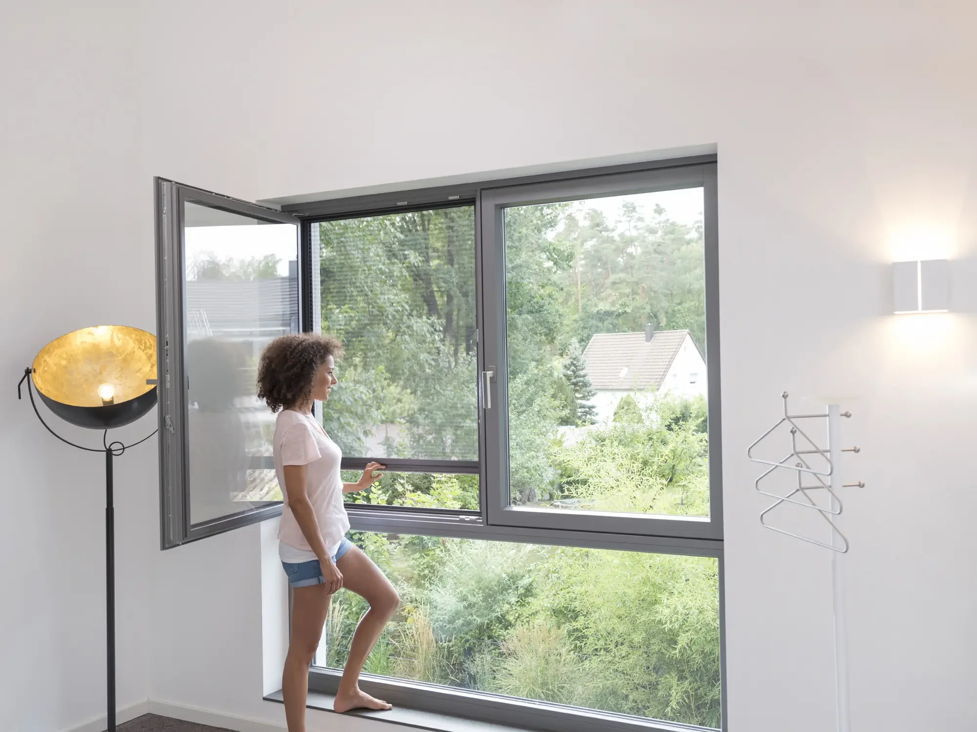 Eine Frau steht vor einem offenen Fenster in einem modernen, hellen Raum. Draußen sieht man Bäume, einen Nachbarhaus und grünen Garten