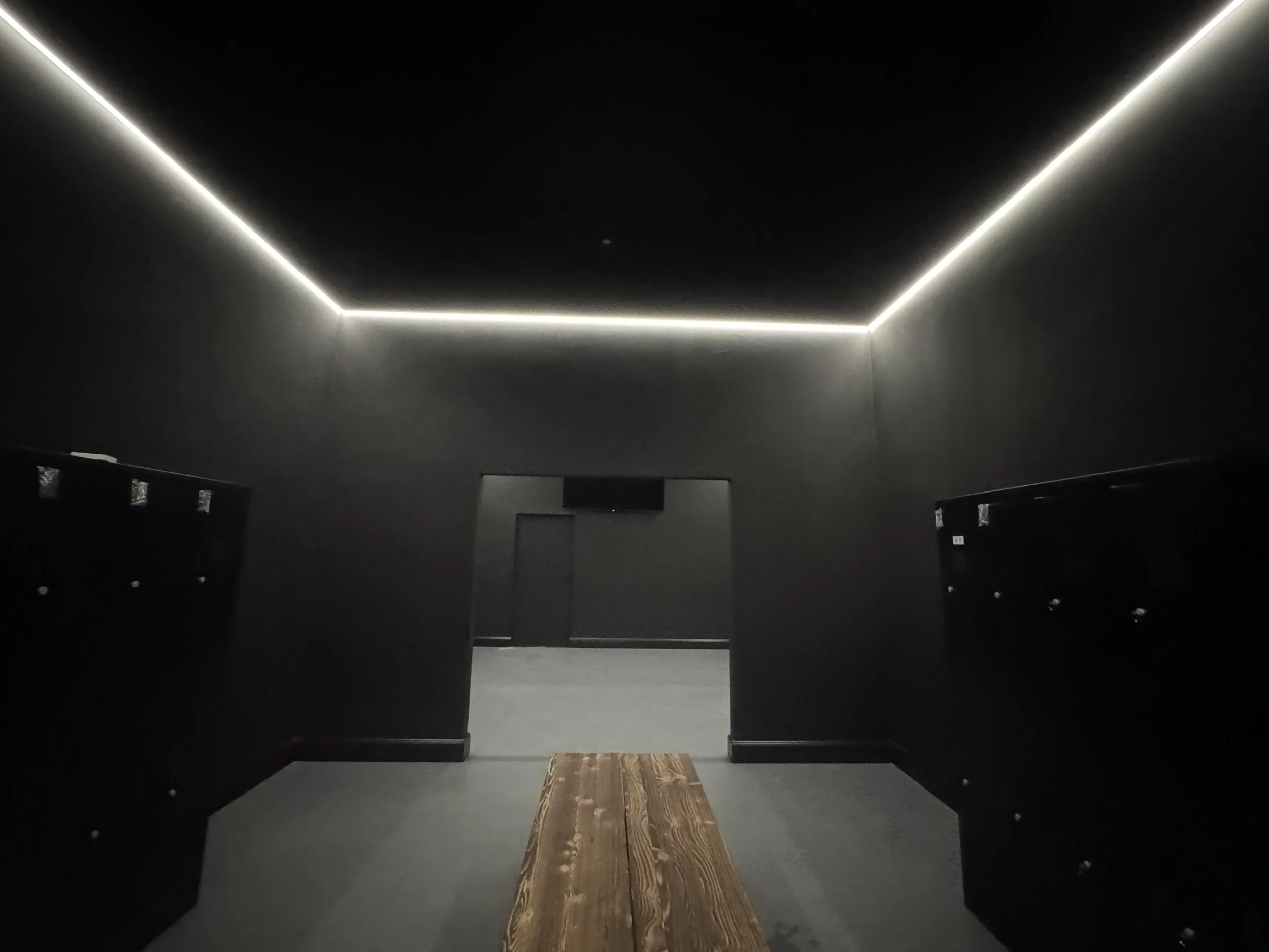 Ein dunkler Raum mit von verstecktem LED-Licht beleuchtetem Decke, die eine rechteckige Form bildet. Im Vordergrund befindet sich ein Holztisch mit einem Teppich darunter, umgeben von schwarzen Stühlen.