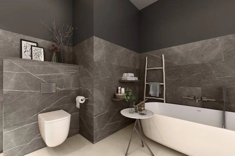 Modernes Badezimmer mit grauen Wandfliesen, freistehender Badewanne, Wandhalterung für Handtücher, kleines Beistelltisch mit Dekor und Pflanzen, weiße Toilette, dunkle obere Wand, dekorative Bilder und Vase mit Zweigen.