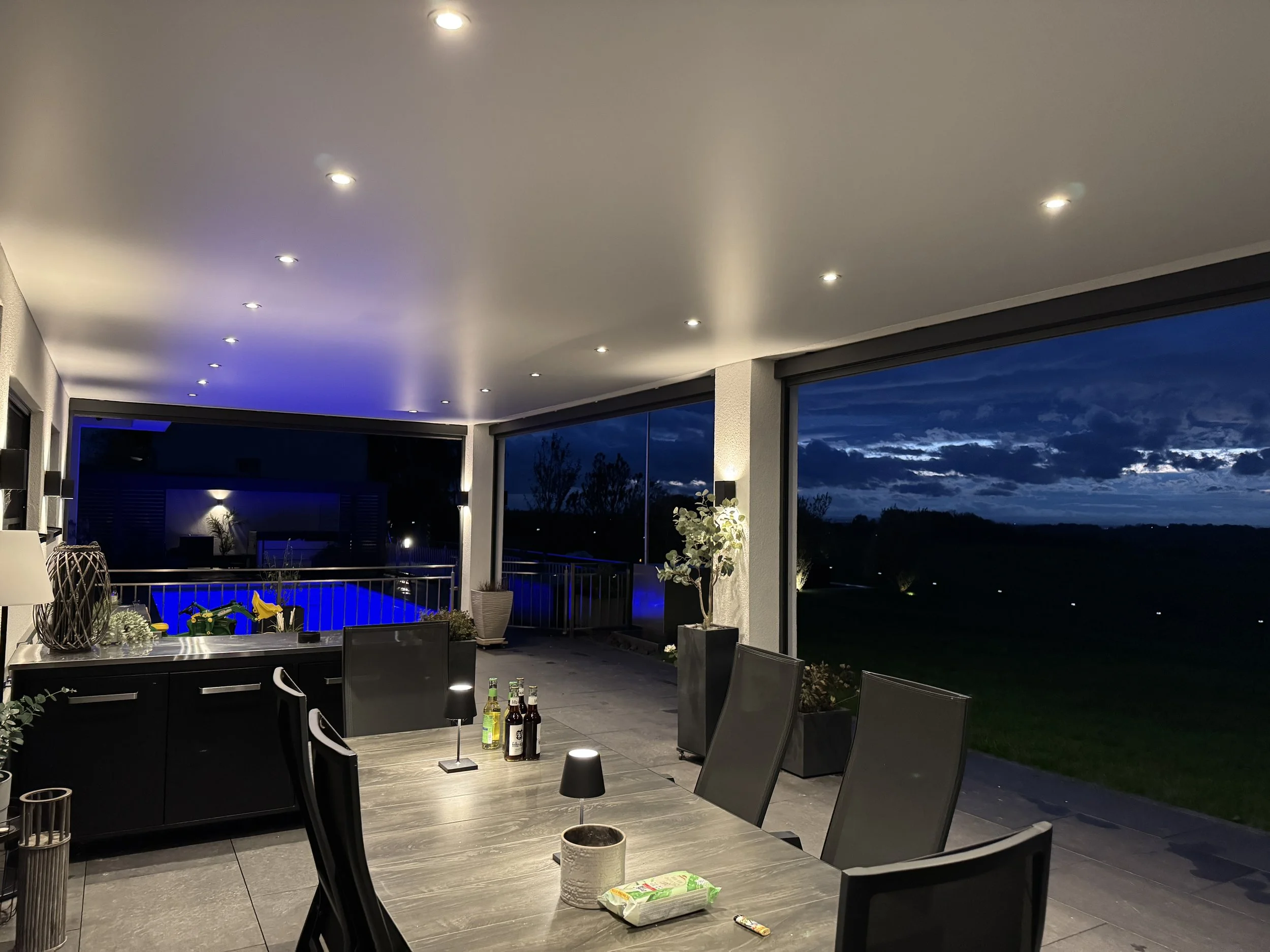 Geräumige Terrasse mit Sitzecke, modernen Möbeln, es gibt Getränke auf dem Tisch, und die Terrassentür führt zu einem dunklen Himmel bei Nacht.