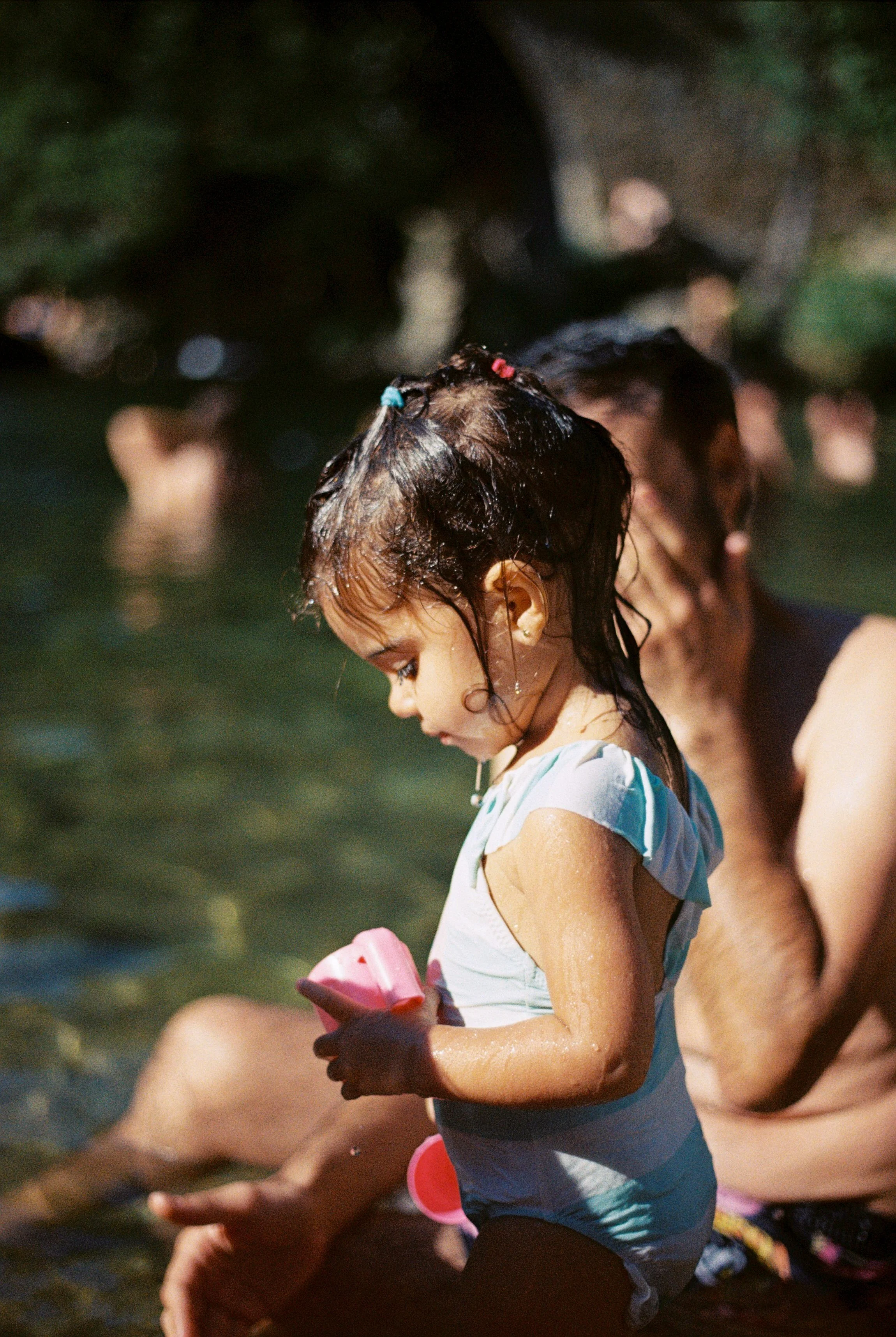 A young girl with wet hair tied with colorful beads, wearing a light striped shirt, holding a pink toy, and standing in a body of water with other children around her, during a sunny day.