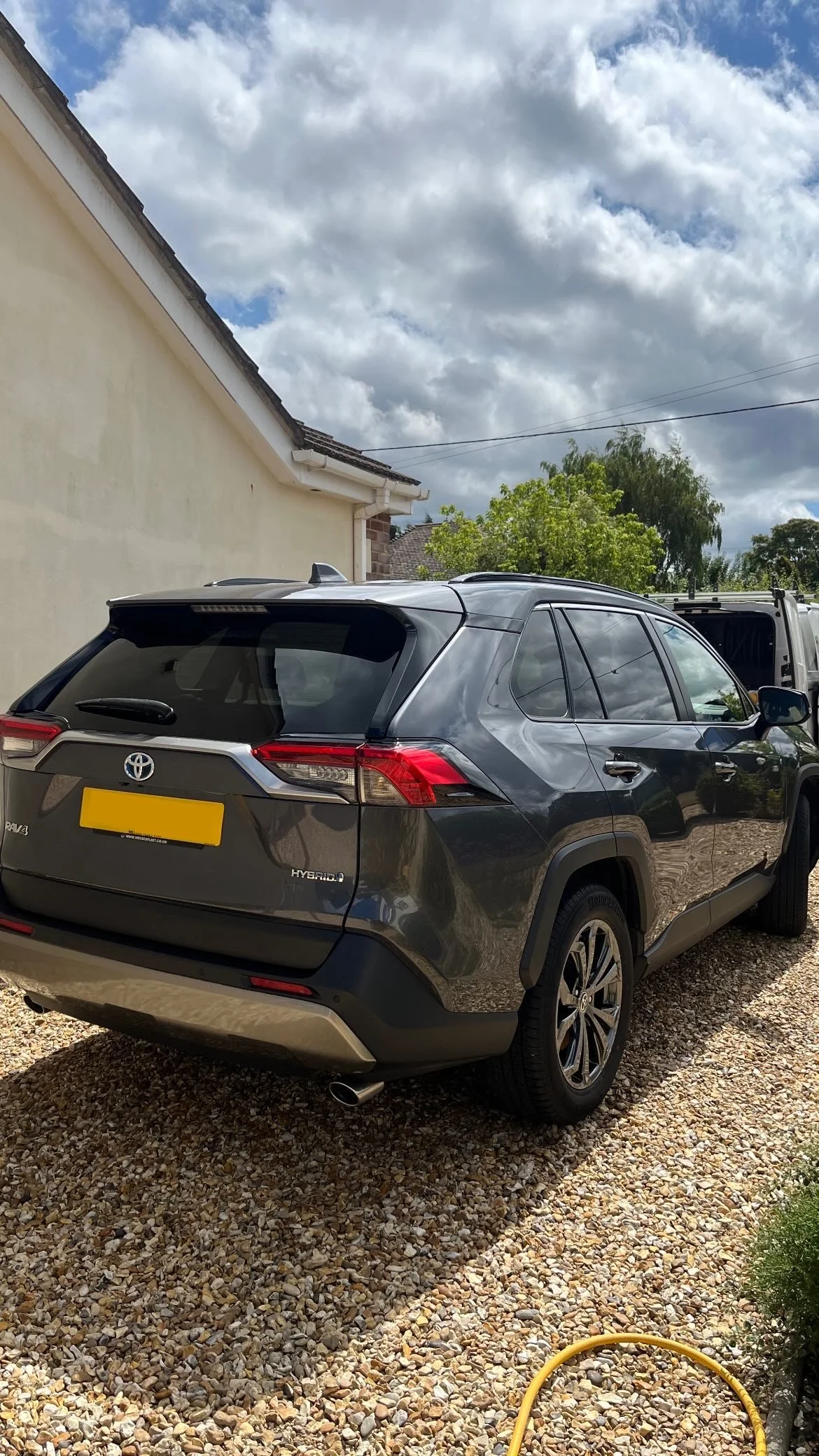 A dark gray Toyota RAV4 Hybrid parked on a gravel driveway next to a house with a yellow wall. The background features a partly cloudy sky, green trees, and a white utility truck.