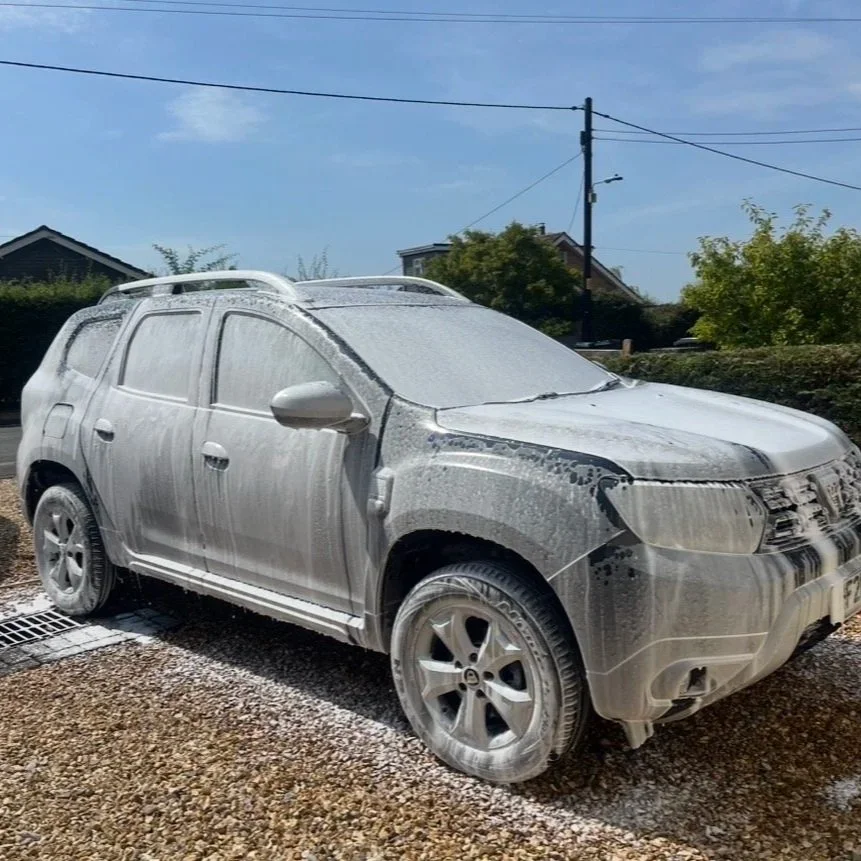 A silver SUV covered in soap and foam during a car wash outdoor on a driveway with a house and trees in the background.