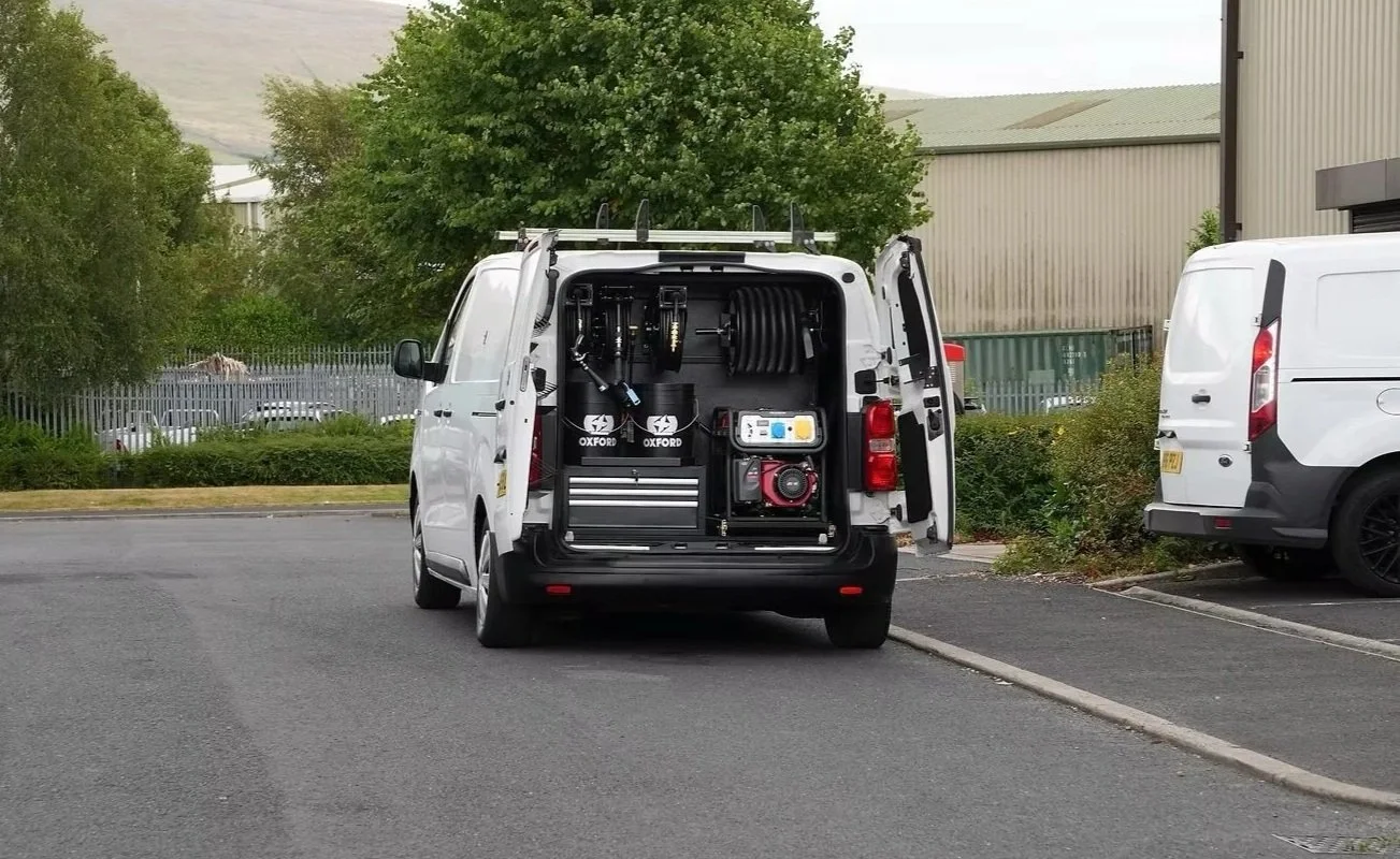 White utility van parked outdoors with its rear doors open, revealing various tools and equipment inside.