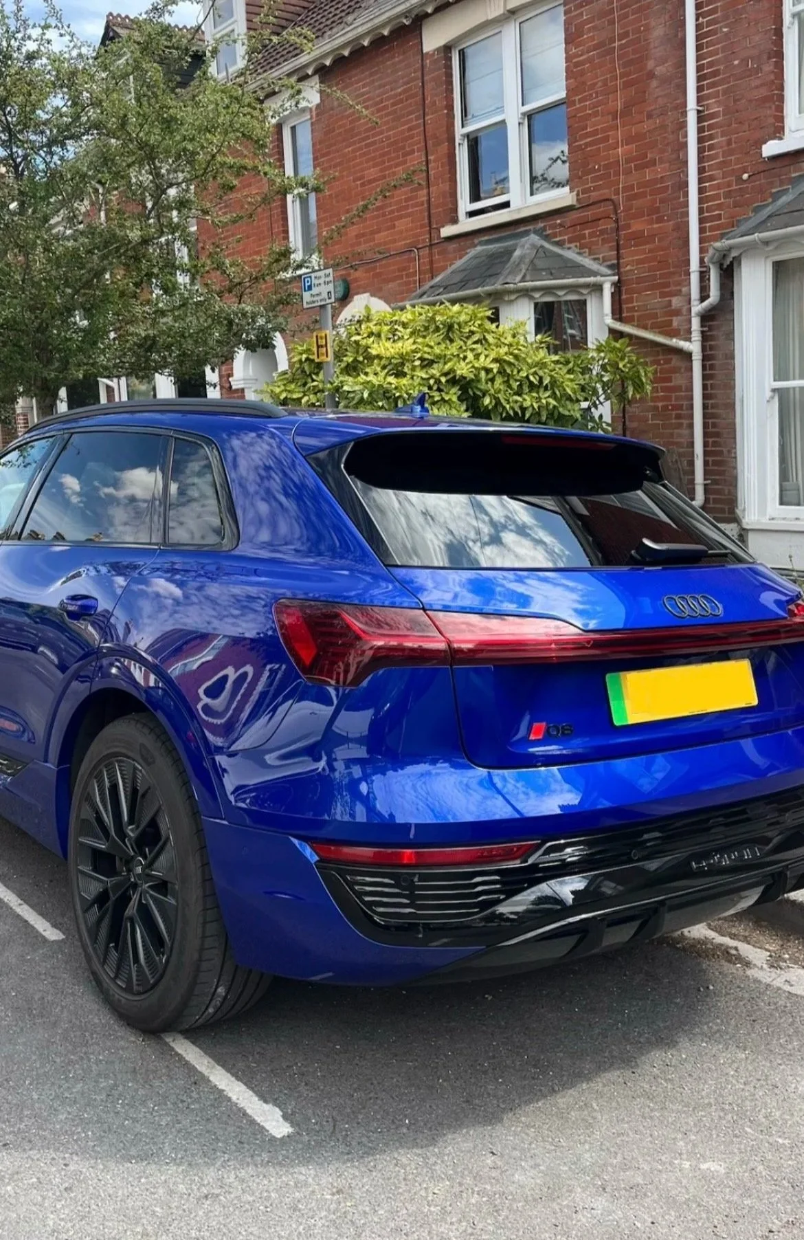 A blue Audi S6 parked in front of a brick house with white window frames, a small bush, and street signs, under a cloudy sky.