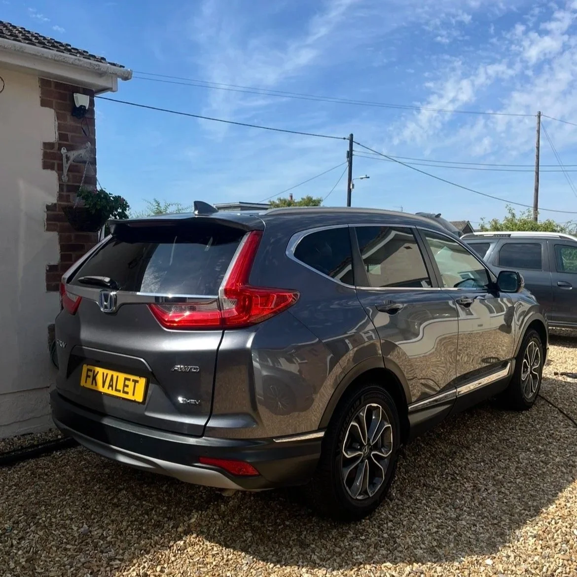 A gray Honda CR-V parked on a gravel driveway outside a house on a sunny day with a blue sky, electrical wires, and other vehicles in the background.