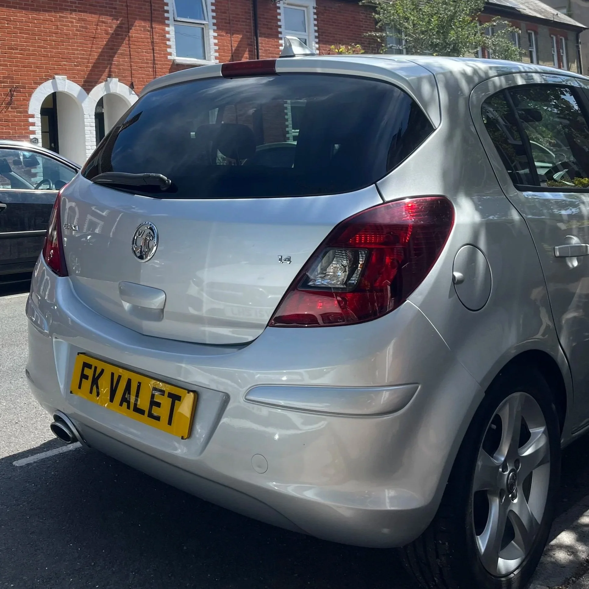 Silver Vauxhall Astra car in a parking lot, with a yellow license plate and a rear windshield wiper, parked near a brick building with white window frames.
