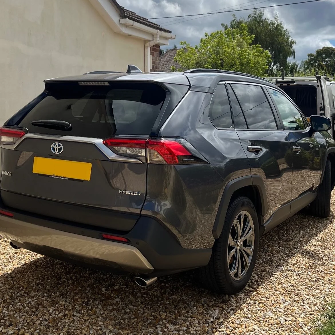 Rear view of a black Toyota RAV4 Hybrid parked on a gravel driveway with trees and a house in the background.