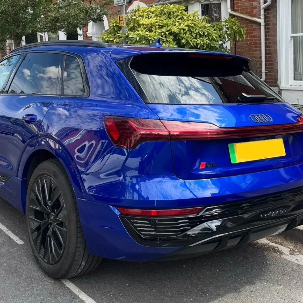 A blue Audi Q6 SUV parked on a street with reflections of the sky and trees on its surface, next to a brick house with a white window and an overgrown bush.
