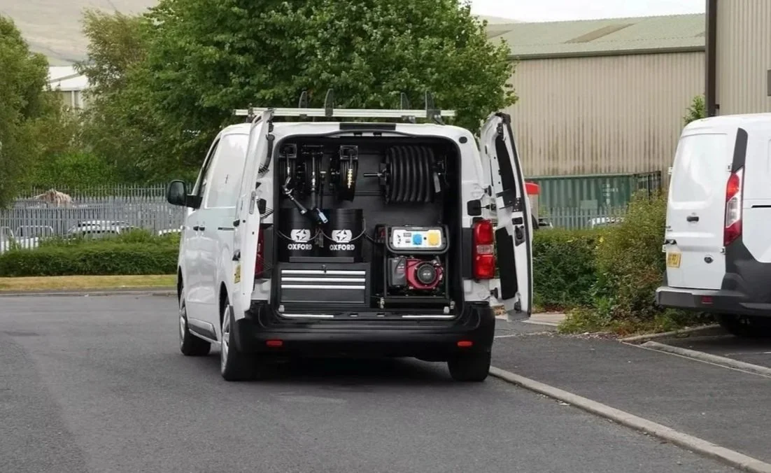 Back of a white utility van with open doors showing equipment inside, including hoses, containers, and tools, parked on a street.