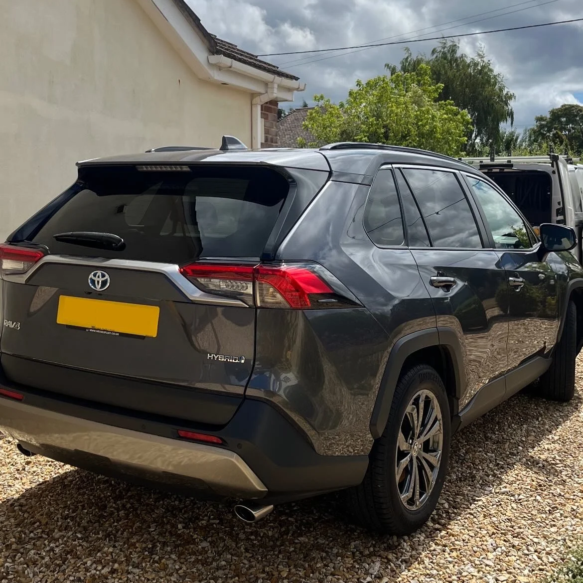 A dark gray Toyota RAV4 Hybrid parked on a gravel driveway next to a beige house with a sloped roof. The rear of the vehicle is visible, showing the Toyota logo, RAV4 and Hybrid badges, and rear lights. There are trees and a cloudy sky in the background.