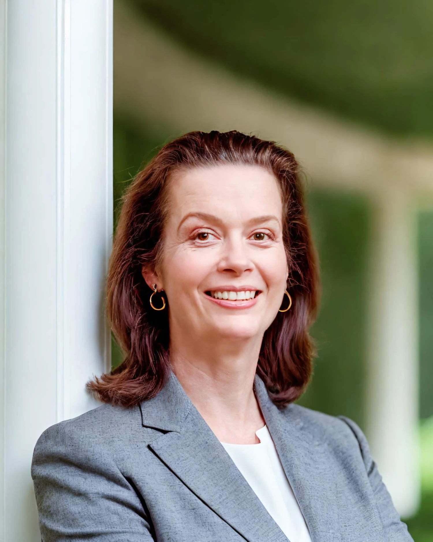 A woman with shoulder-length auburn hair smiling, standing outside near a white column, wearing a gray blazer and earrings.