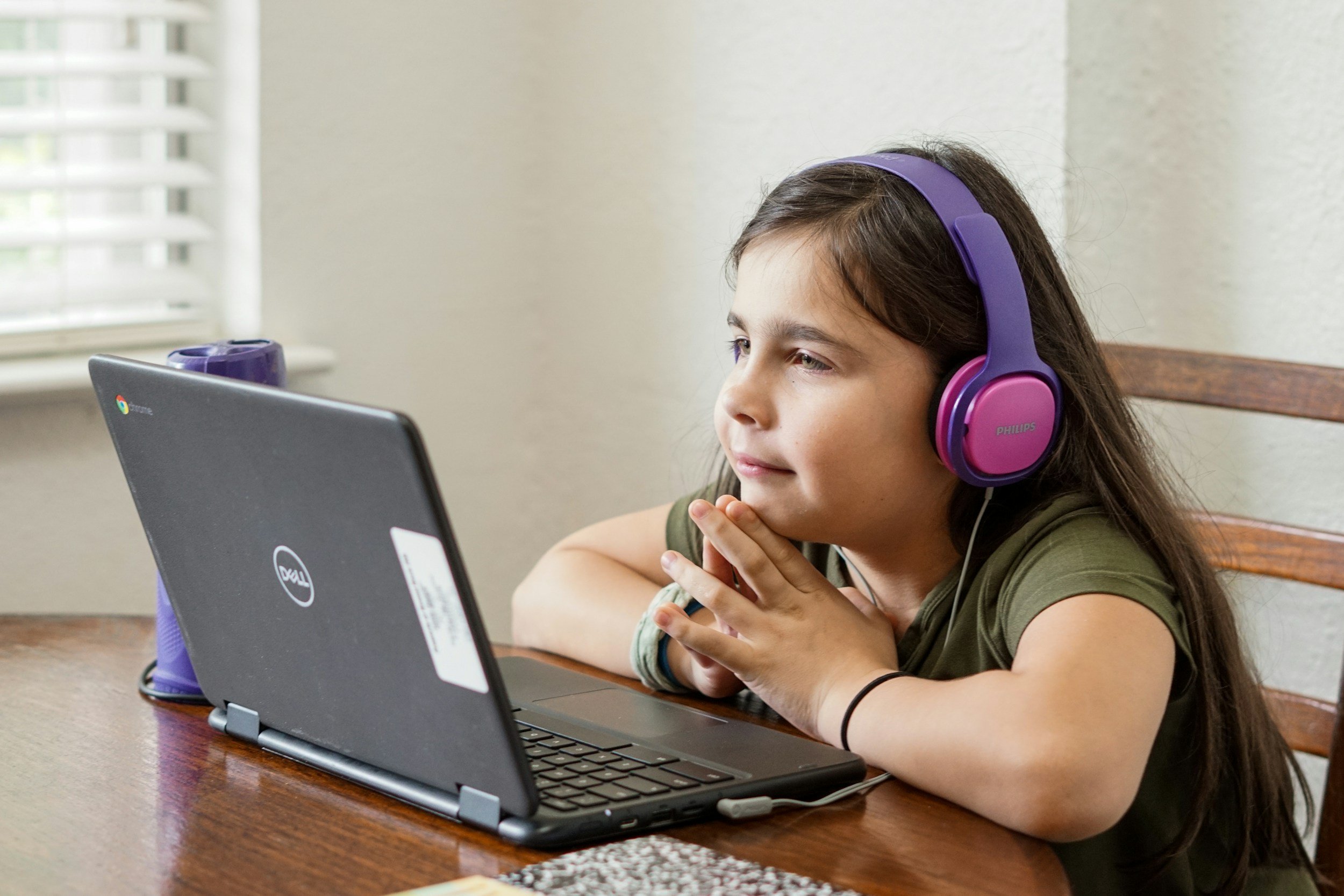 A young girl with long hair wearing pink and purple headphones sitting at a wooden table with her chin resting on her hands, looking at a laptop screen.