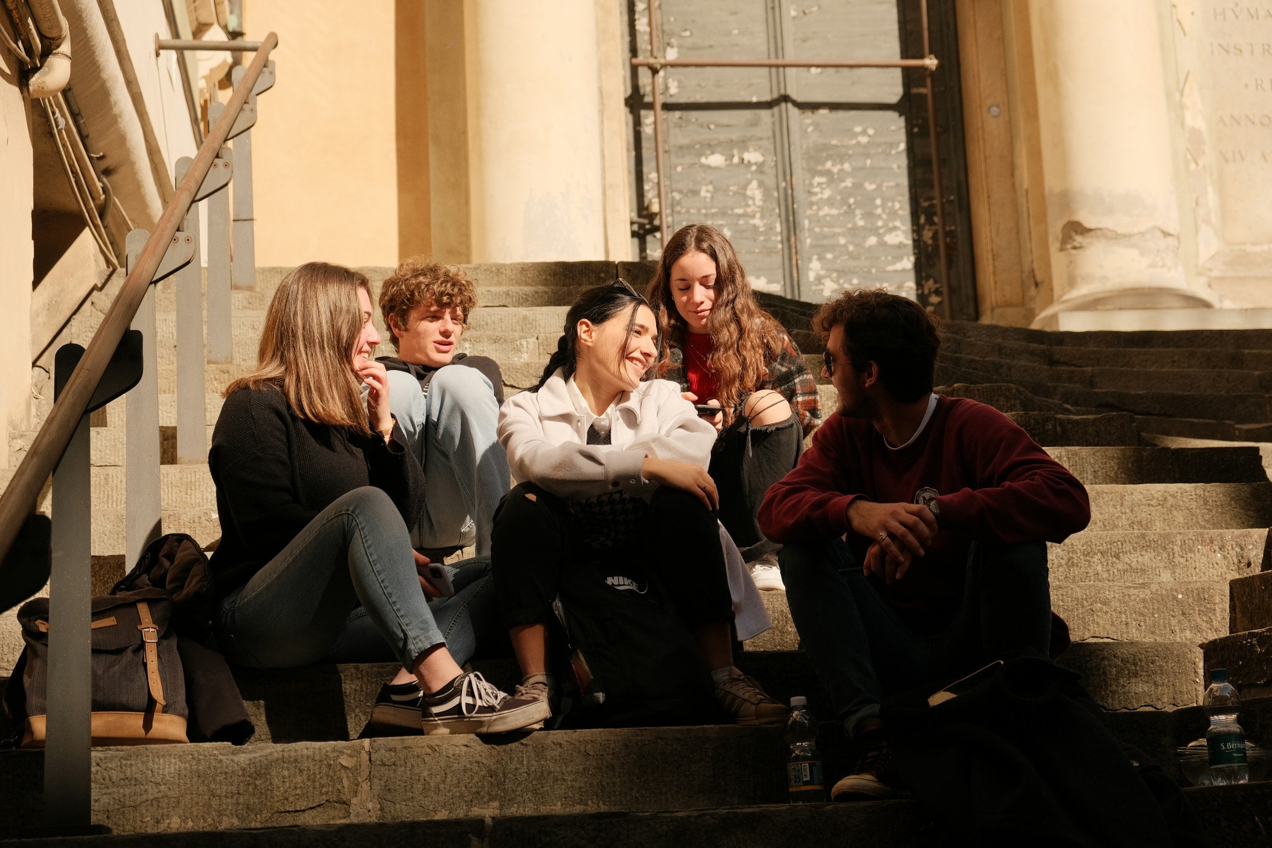Group of five young people sitting on stone steps outside, engaging in conversation and smiling, in front of a historic building with columns.