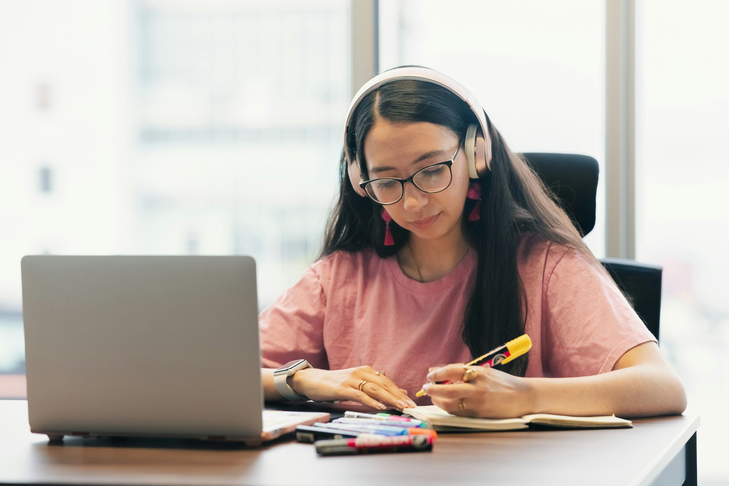 Young woman with long dark hair, glasses, and pink earrings, wearing a pink T-shirt, working at a desk with a laptop, colorful markers, and paper, in a bright office environment.