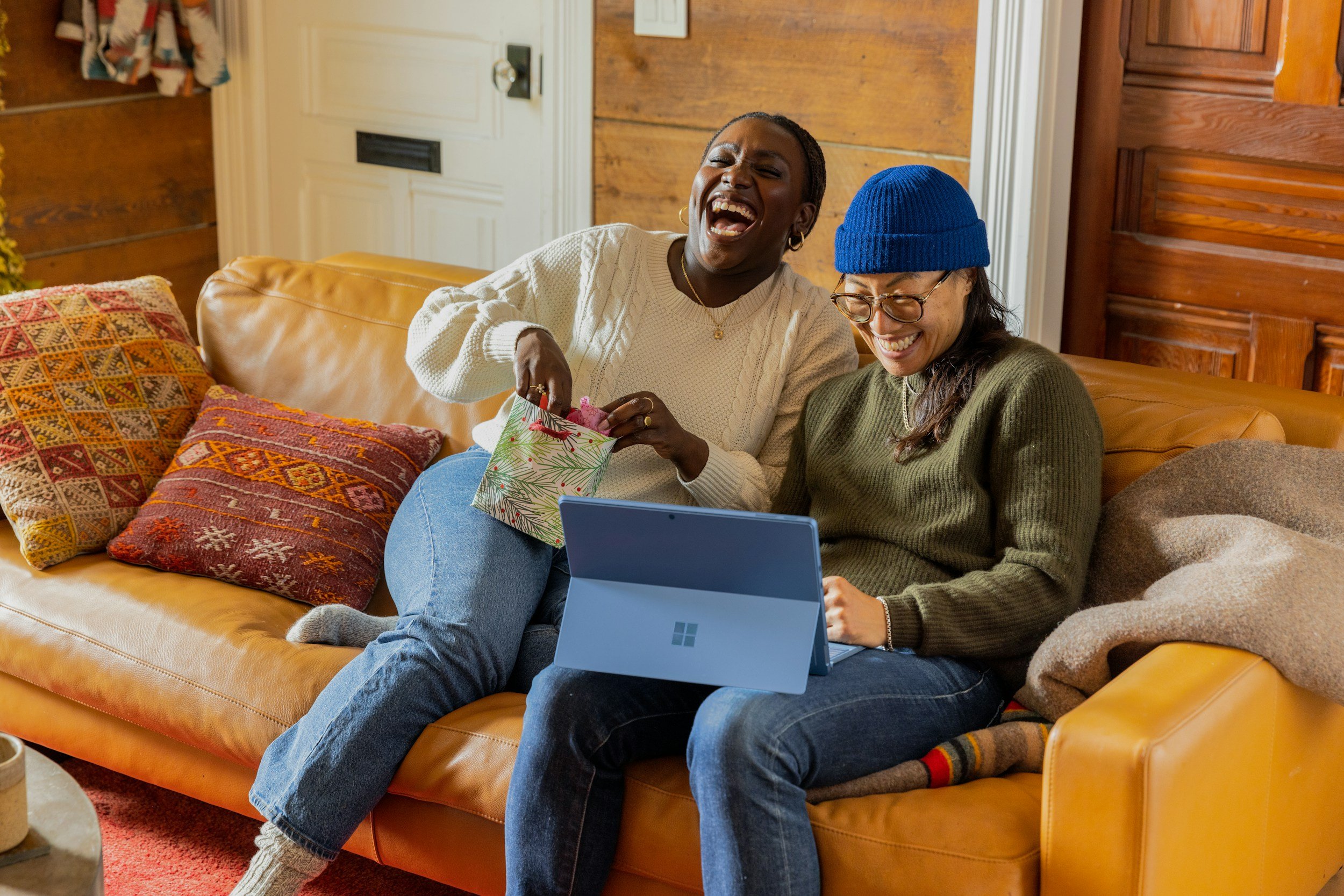 Two women sitting on a yellow leather couch, laughing, with one holding a gift bag and the other using a laptop. They are in a cozy room decorated with patterned pillows, wooden walls, and a soft carpet.