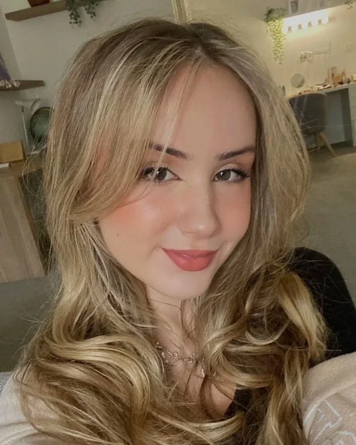 A young woman with long, blonde, wavy hair smiling at the camera in a cozy indoor setting.
