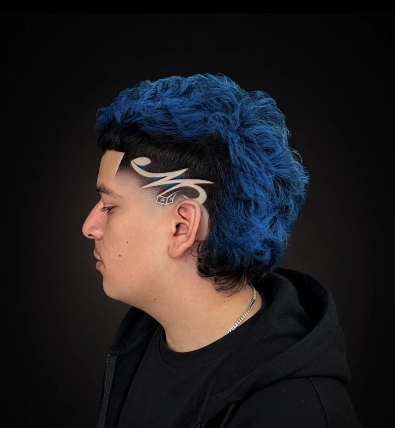 Side profile of a young man with black hair and bright blue curly hair on top, featuring a lightning bolt and racing car painted on the side of his head against a black background.
