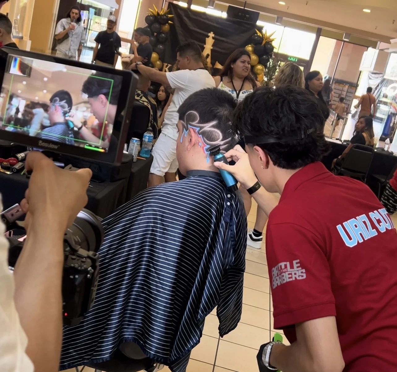 A young man is getting a colorful face paint design in a public indoor space, with a person in a red shirt assisting. There is a camera capturing the moment, and several onlookers and staff are visible in the background, with decorations and black and gold balloons.