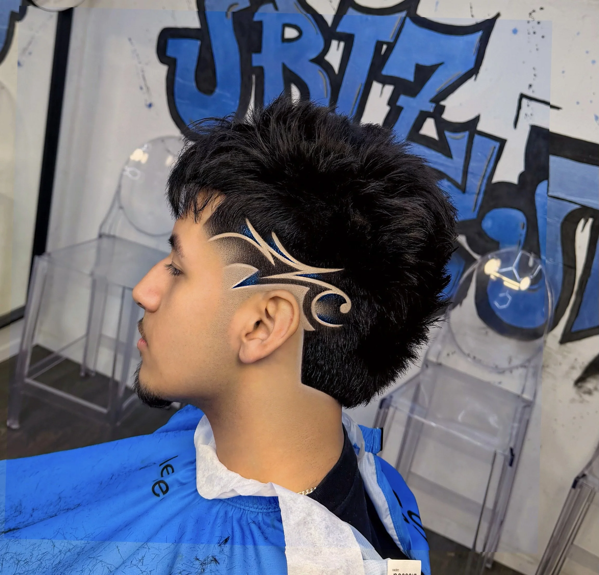 A person with a short, styled haircut featuring intricate black and beige face paint designs, wearing a blue apron, standing in front of a graffiti-style art background.