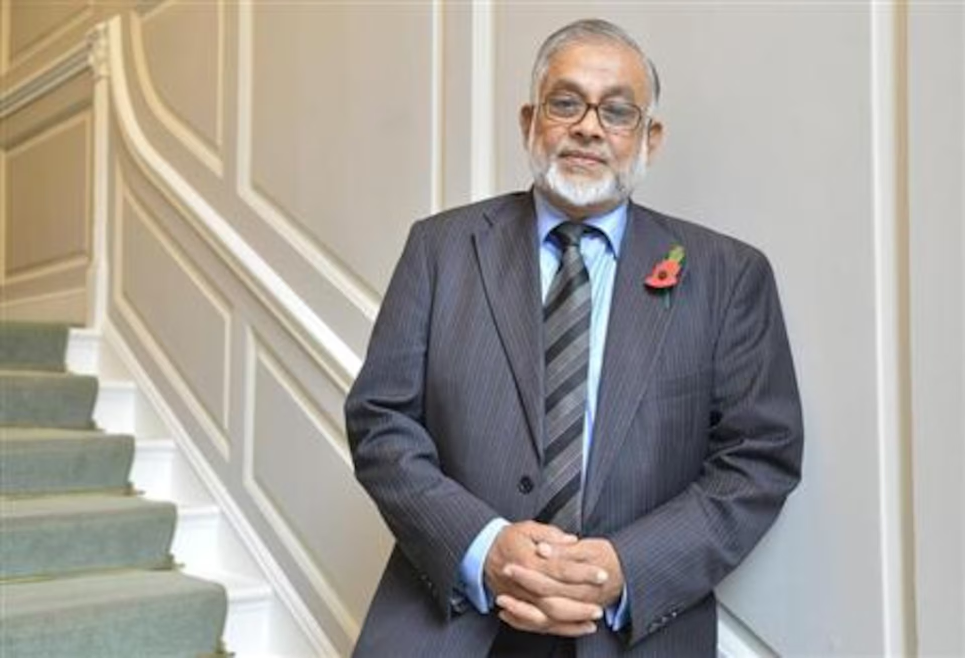 An elderly man dressed in a dark pinstripe suit, light blue shirt, and a striped tie, standing indoors near a staircase with beige walls and white trim. He has gray hair, glasses, and a white beard, with a red poppy pinned to his suit lapel, and he's holding his hands together in front of him.