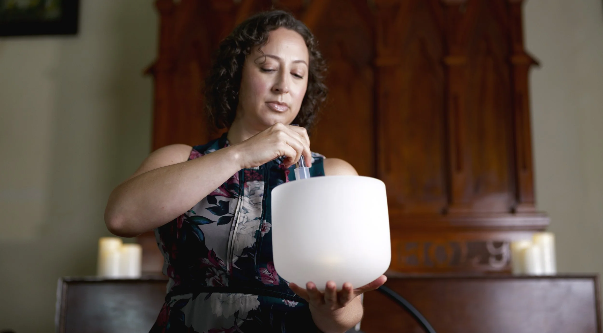 A woman with curly hair in a sleeveless floral dress looks down as she plays a white crystal singing bowl with a mallet.