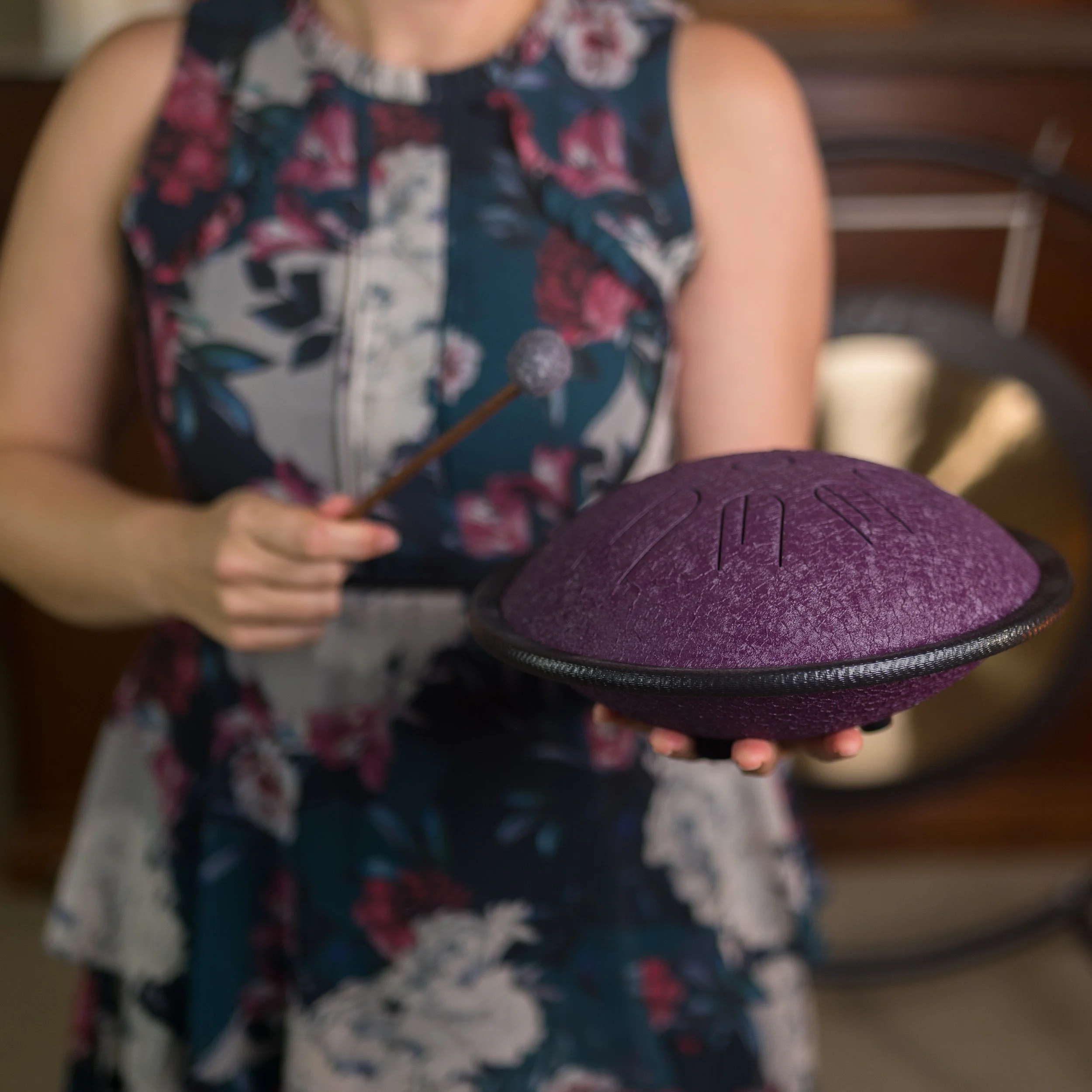 A woman wearing a sleeveless floral dress holding a purple tongue drum and mallet.