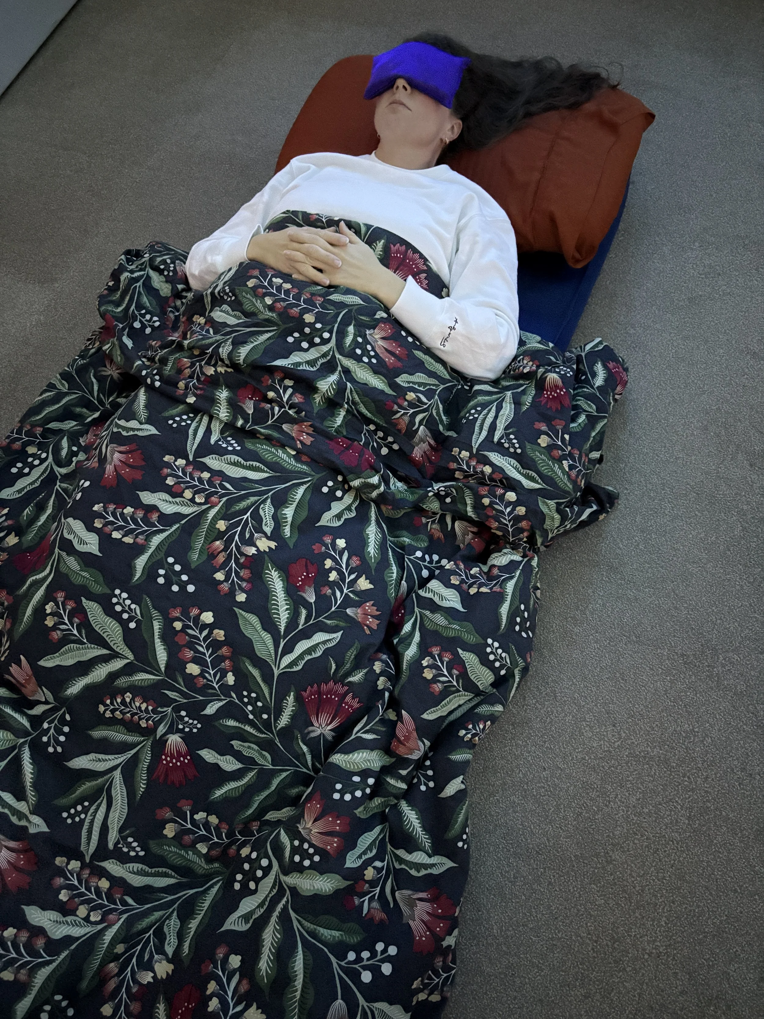 A woman lying on her back with a violet eye mask, her head on an orange pillow and a floral blanket over her.