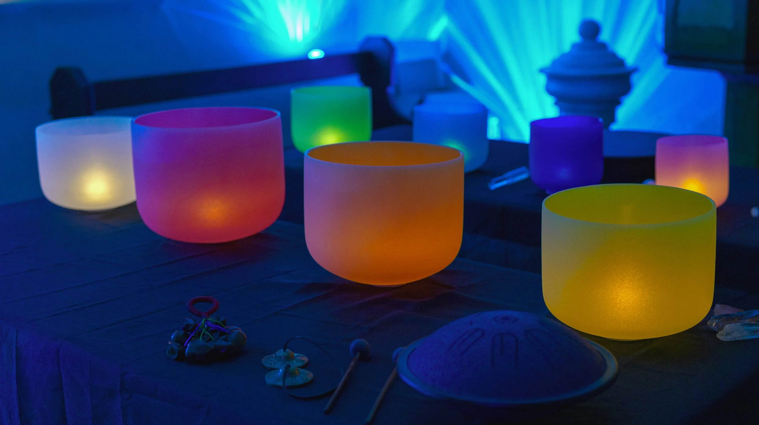 Coloured crystal singing bowls lit by tea lights against a glow of green and blue, with a tongue drum, tingsha bells and a seed pod rattle in front.