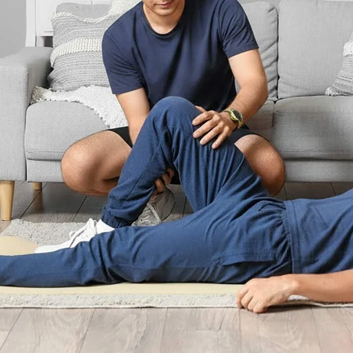 A person is lying on the floor with their arm resting on the ground, while another person kneels next to them, holding their leg in a physical therapy or medical assistance setting.