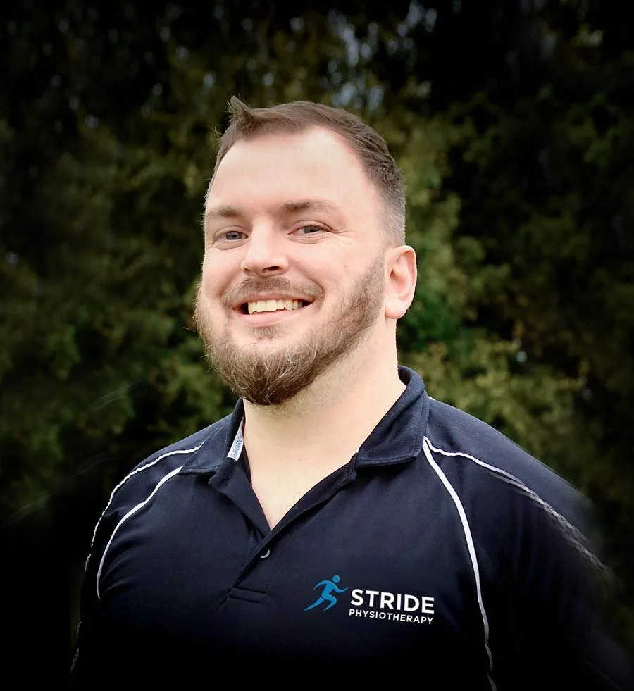 Smiling man with short hair and a beard wearing a black sports polo shirt with white piping and a 'Stride Physiotherapy' logo, standing outdoors with trees in the background.