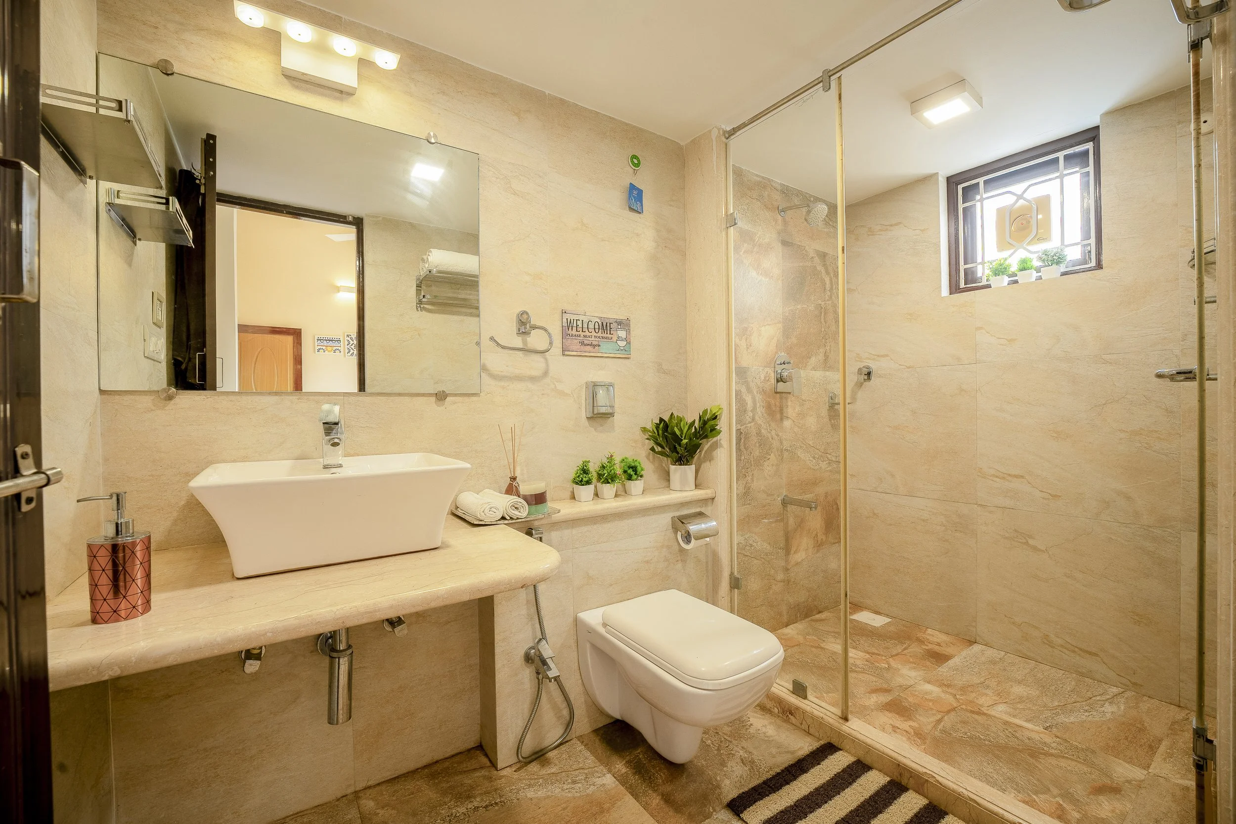 3BHK Villa In Goa With a Bathroom with a vanity, mirror, towels, plants, toilet, and a shower with a window showing potted plants outside.