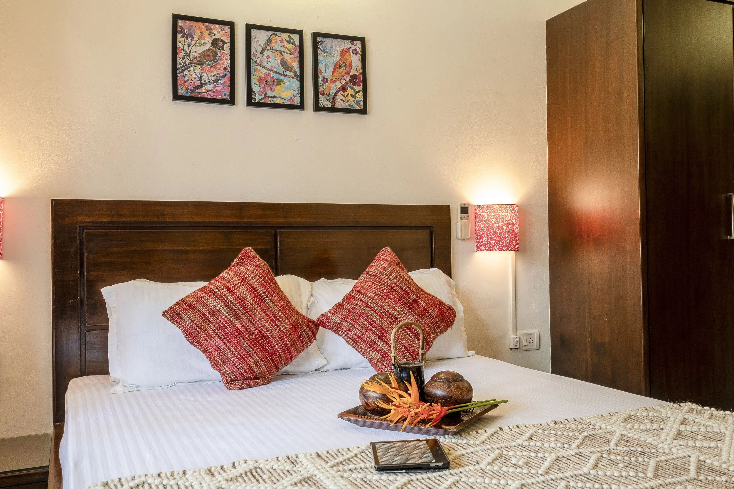 A bedroom with a wooden headboard and two pillows with red patterned covers. There are two lamps with red patterned shades on both sides of the bed. Above the bed, there are three framed bird-themed artworks. A tray with tropical decorations and a smartphone are on the bed.