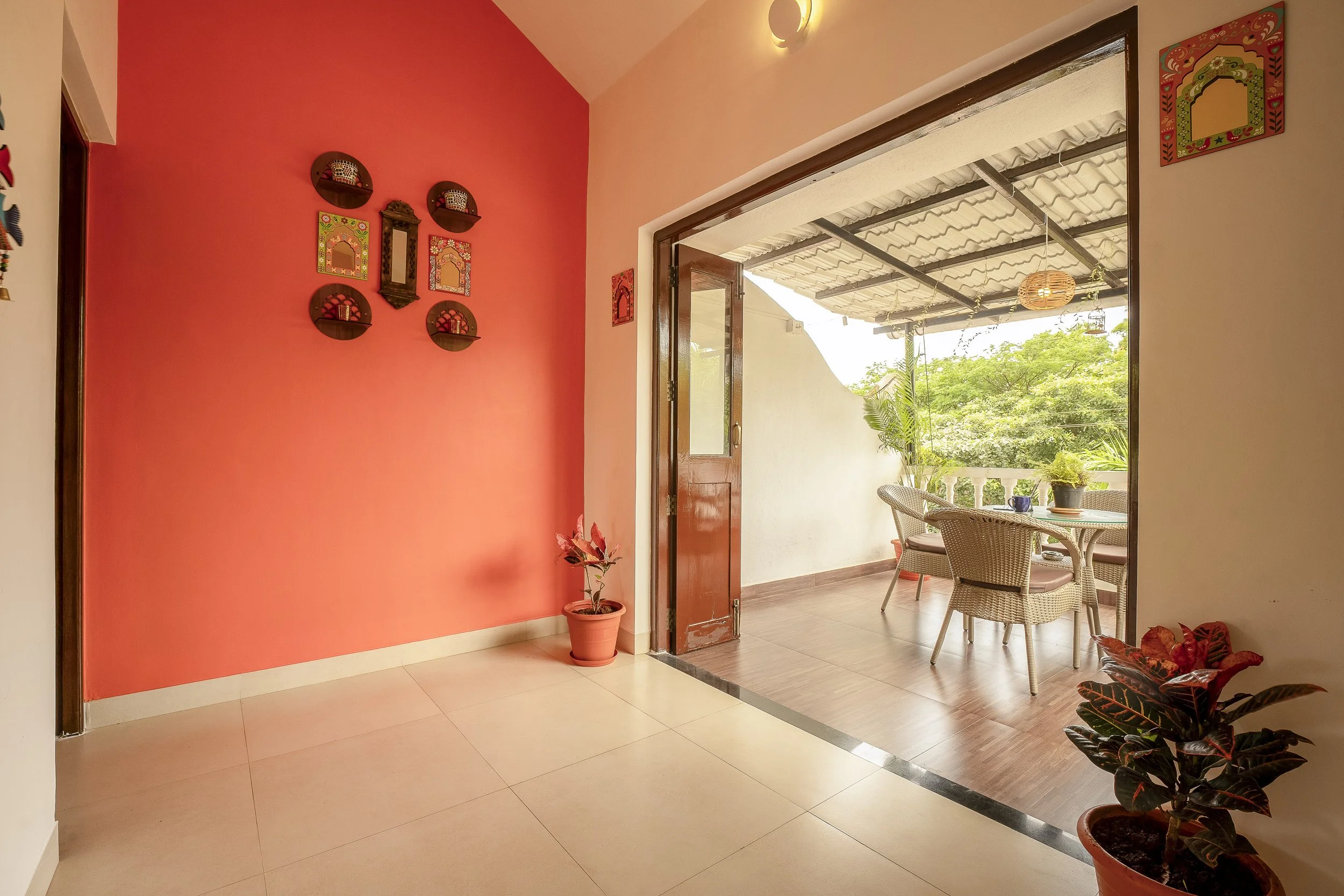 3BHK Villa In Goa With A bright indoor patio with a pink accent wall, decorated with small shelves and colorful framed artwork. There are potted plants on the floor and on the table, and an open wooden door leading to an outdoor area with lush greene