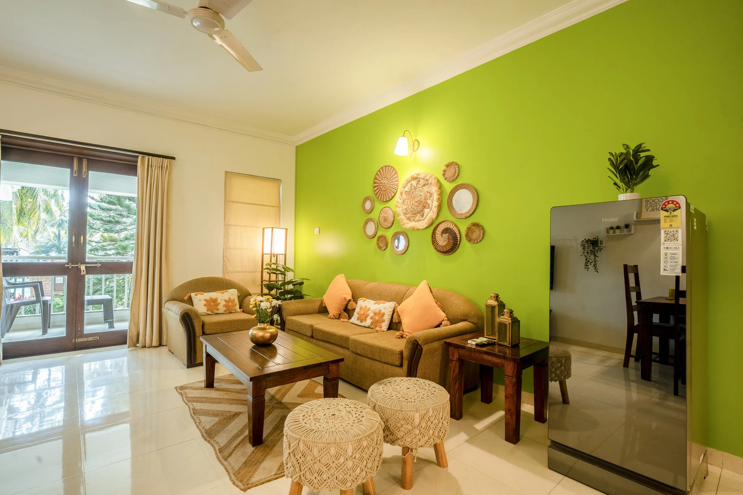 Living room with brown sofa, decorative pillows, a wooden coffee table with a vase, a green accent wall with woven wall hangings, beige curtains, sliding glass door with a balcony, and a corner with a refrigerator and plant.