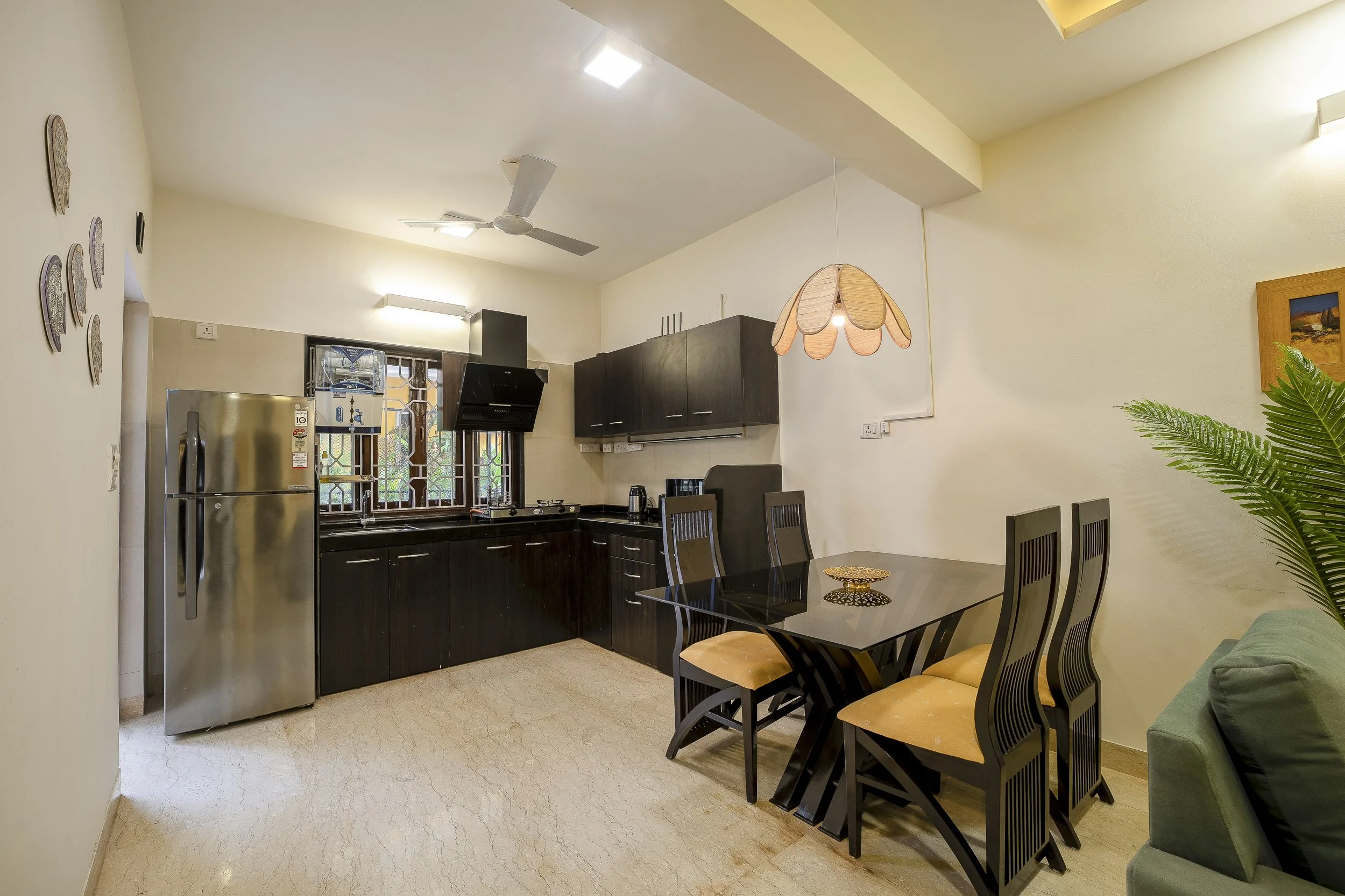 3BHK Villa In Goa With a View of a kitchen and dining area with black cabinets, a stainless steel refrigerator, a dining table with six chairs, hanging light fixture, ceiling fan, and a window with decorative grilles.