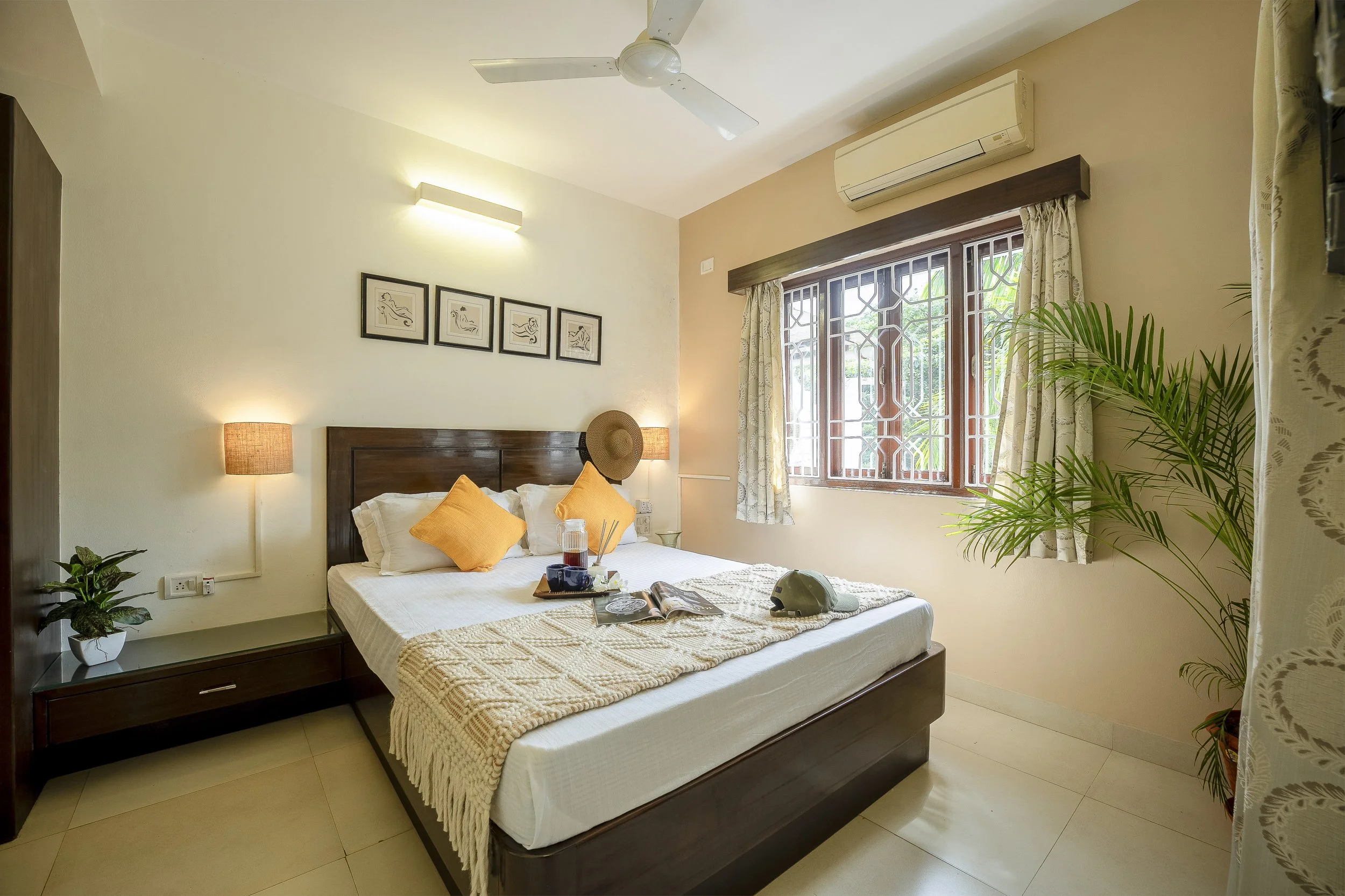 3BHK Villa In Goa with a large bed featuring white bedding, orange pillows, and a beige throw. There are wooden nightstands with lamps, a potted plant, and a ceiling fan. The window has patterned curtains and a view of greenery outside.