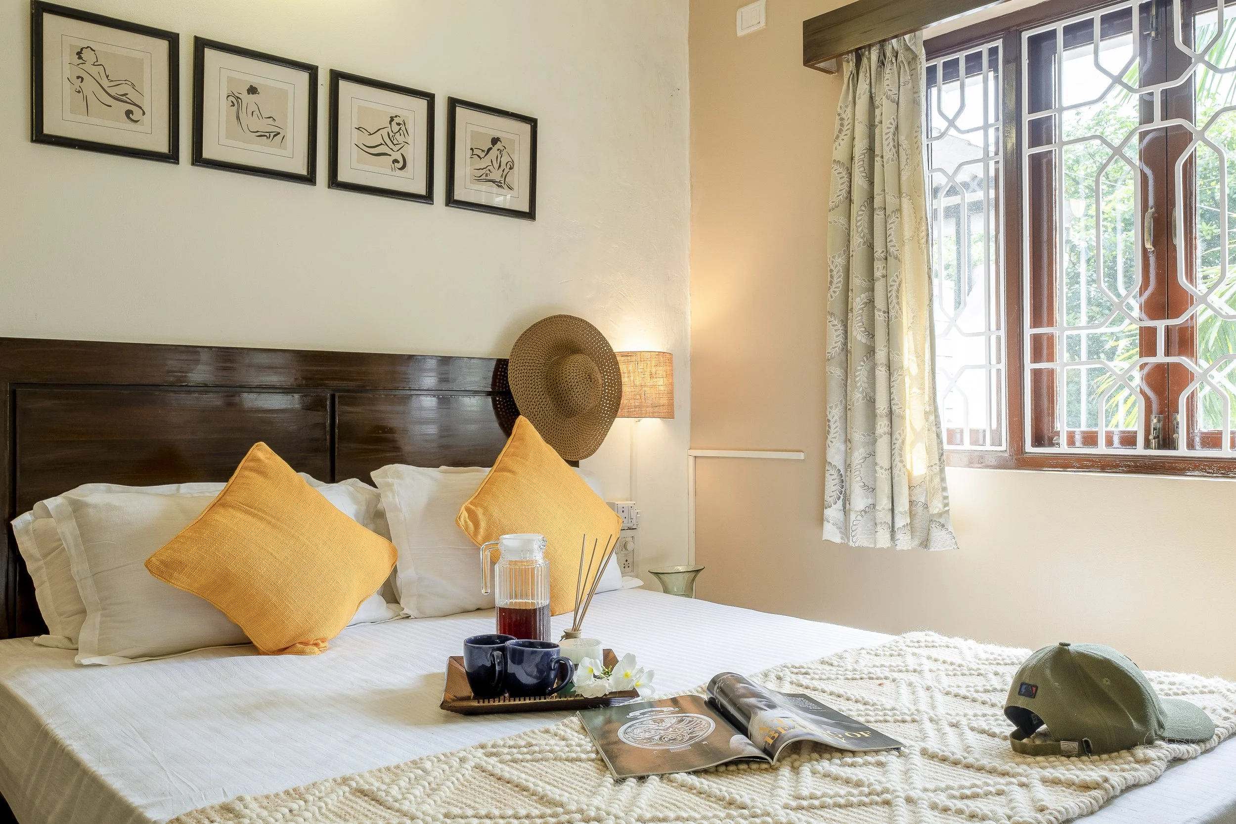 3BHK Villa In Goa With A cozy bedroom with a bed having white sheets and yellow pillows, a wooden headboard, a bedside lamp, framed artwork on the wall, a window with patterned curtains, a tray with a pitcher, cups, flowers, and a magazine on the bed