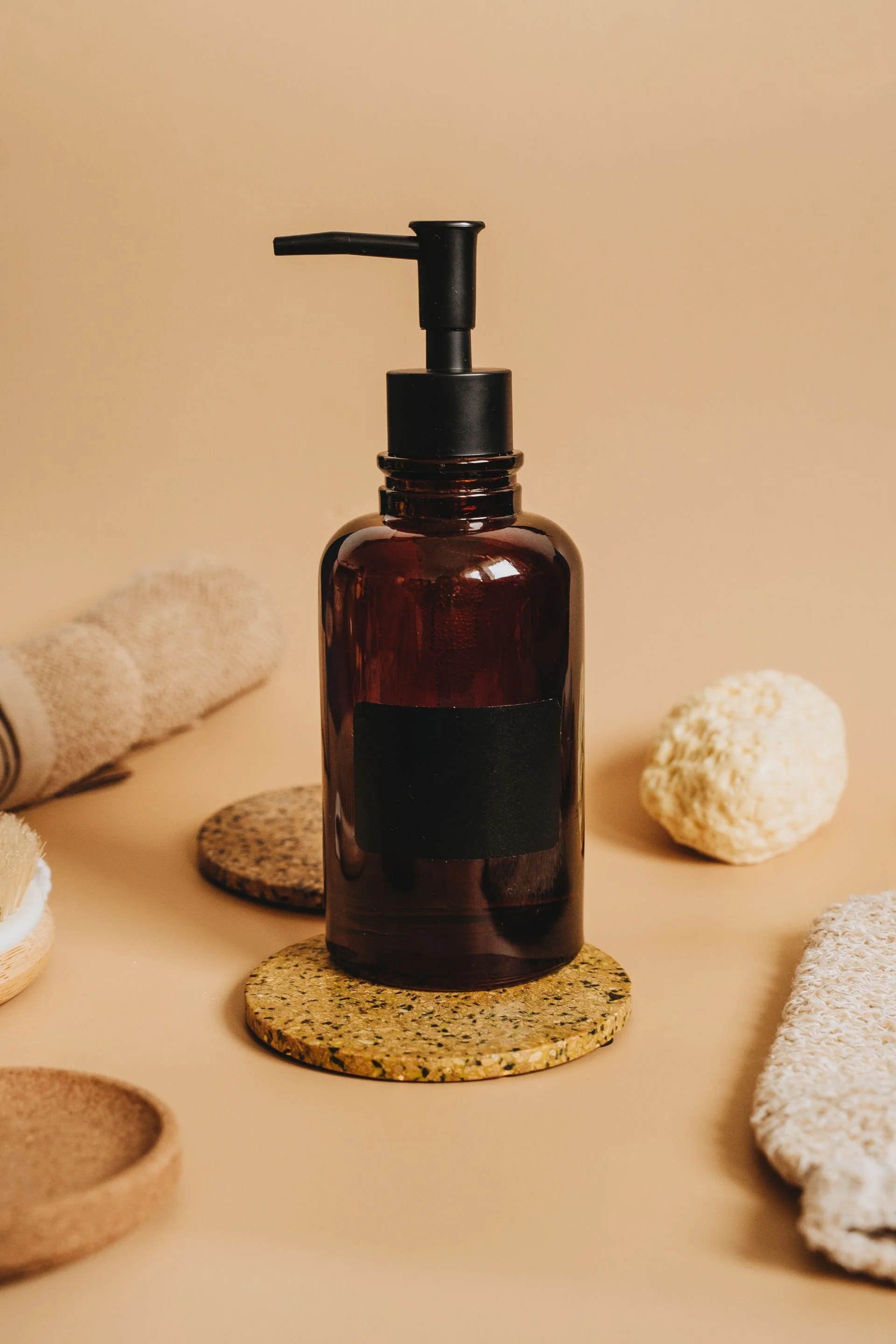 A dark amber glass bottle with a black pump dispenser, placed on a round speckled coaster, surrounded by various stones and textured objects, on a beige background.