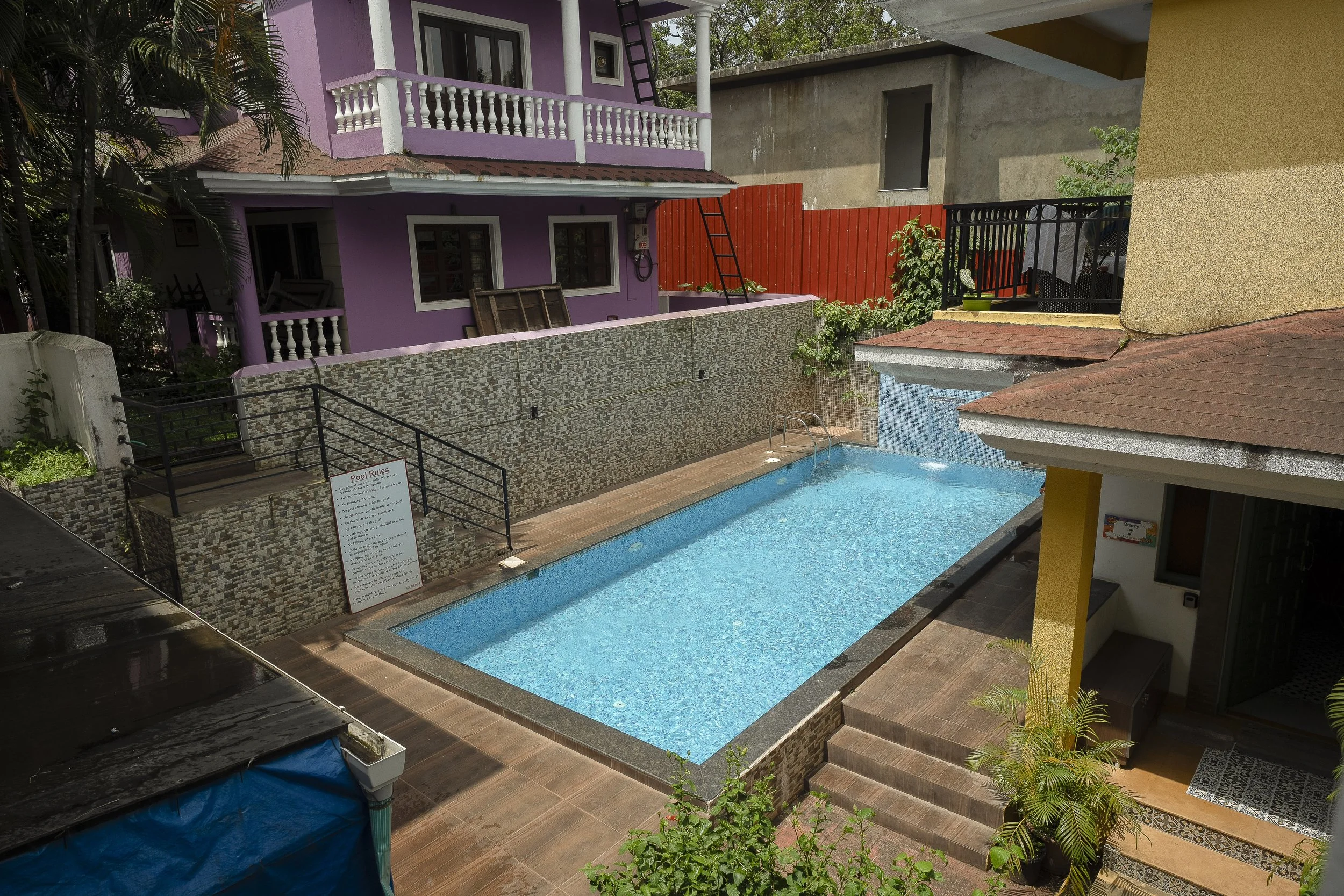 3BHK Villa In Goa With A swimming pool area with a small staircase leading into the pool, surrounded by various buildings including a purple house with white balcony railings, a yellow building, and a partially constructed gray building. There are pl