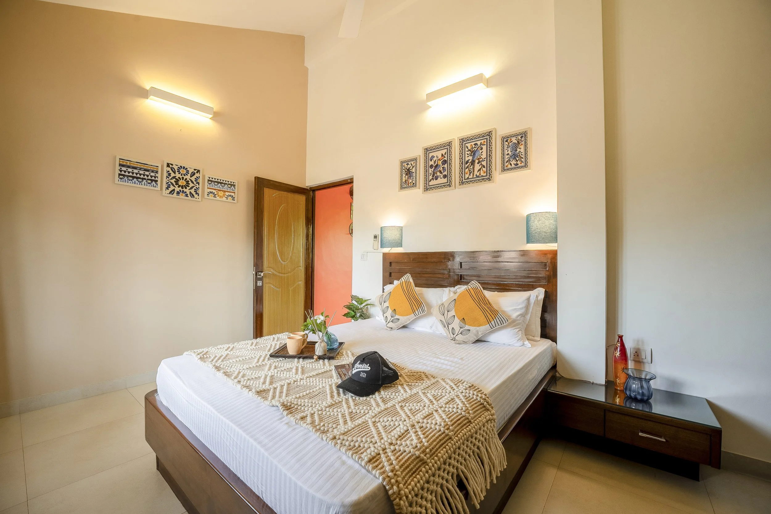 3BHK Villa In Goa With a room with a bed, pillows with a leaf pattern, a nightstand with a blue vase, a lamp, and framed floral artwork on the wall.