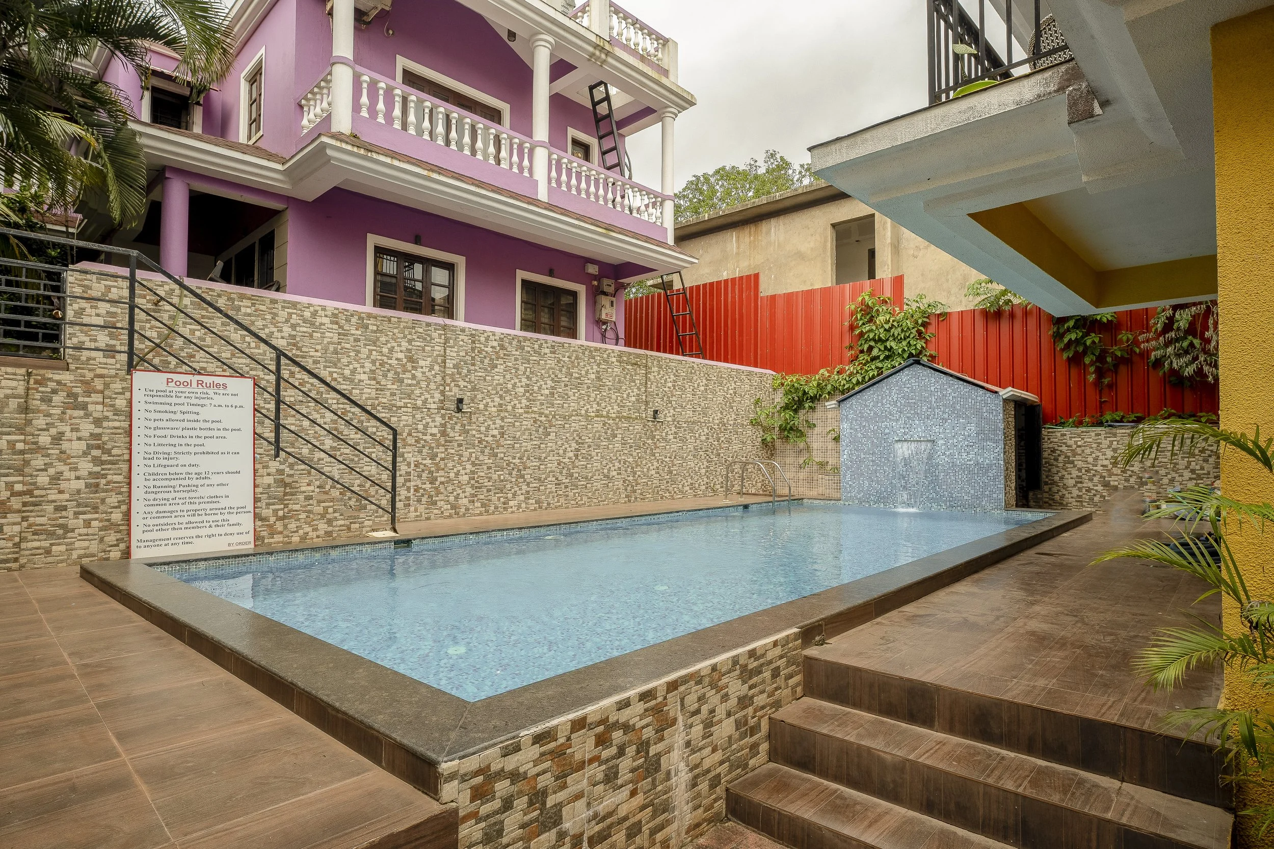 3BHK Villa In Goa With a Swimming pool area surrounded by colorful buildings with purple, yellow, and beige walls. The pool is tiled with small blue mosaic tiles and has a metal ladder. There are steps leading to the pool, and a sign with pool rules 