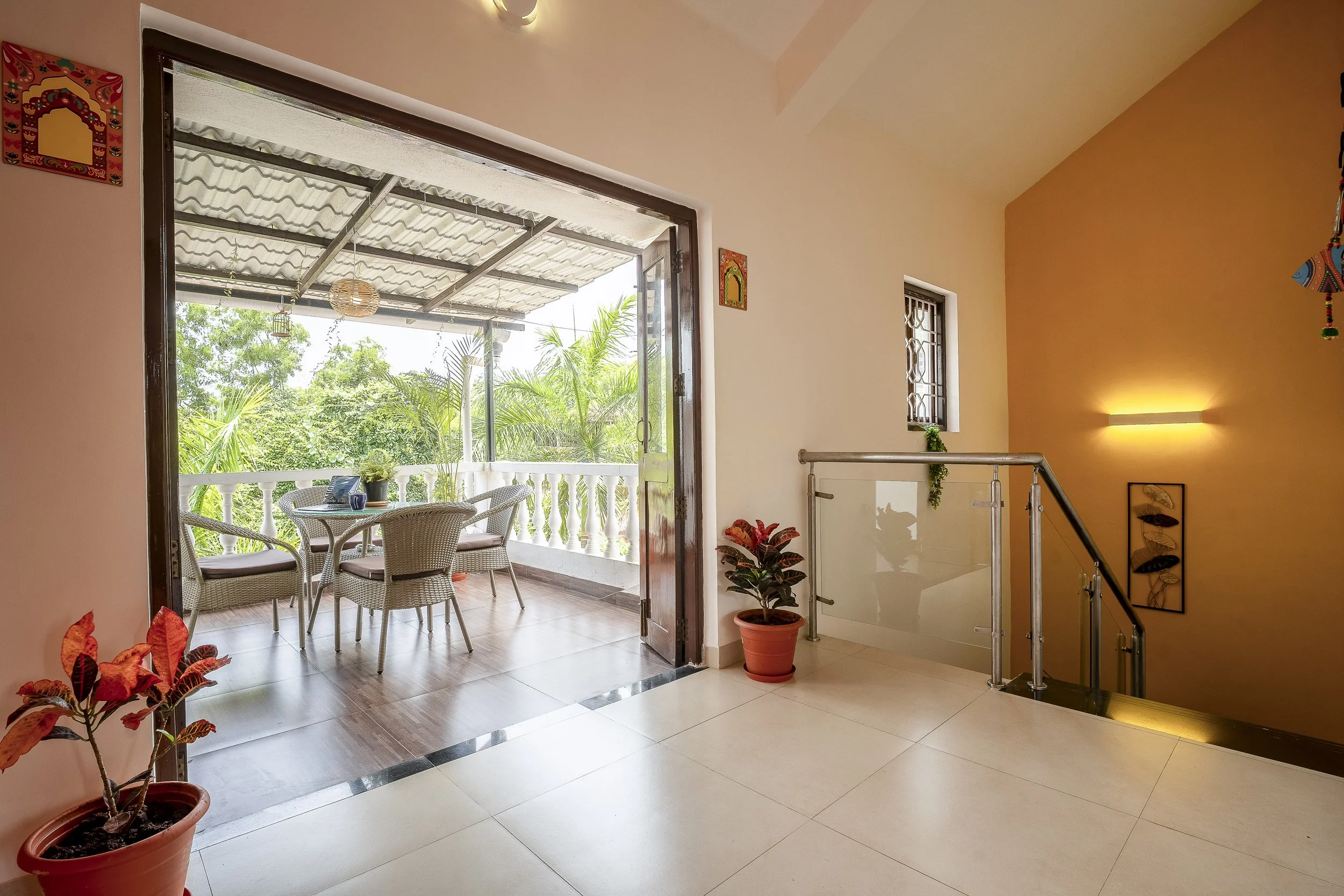 3BHK Villa In Goa With a view of a cozy balcony with patio furniture, surrounded by lush greenery, accessible through an open sliding door from a bright interior with orange accent wall, indoor plants, and wall decorations.