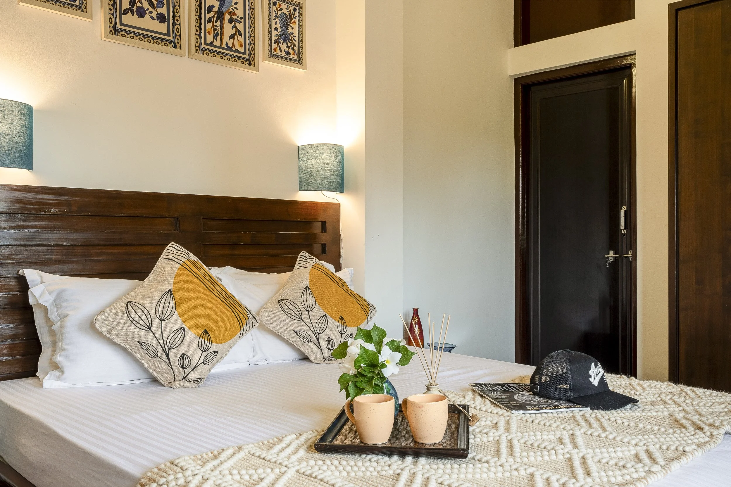3BHK Villa In Goa With a with a bed, beige walls, decorative pillows with leaf patterns, a wooden headboard, wall-mounted blue lamps, a small tray with two pink cups, a floral arrangement, a magazine, a black baseball cap, and a dark-colored door.